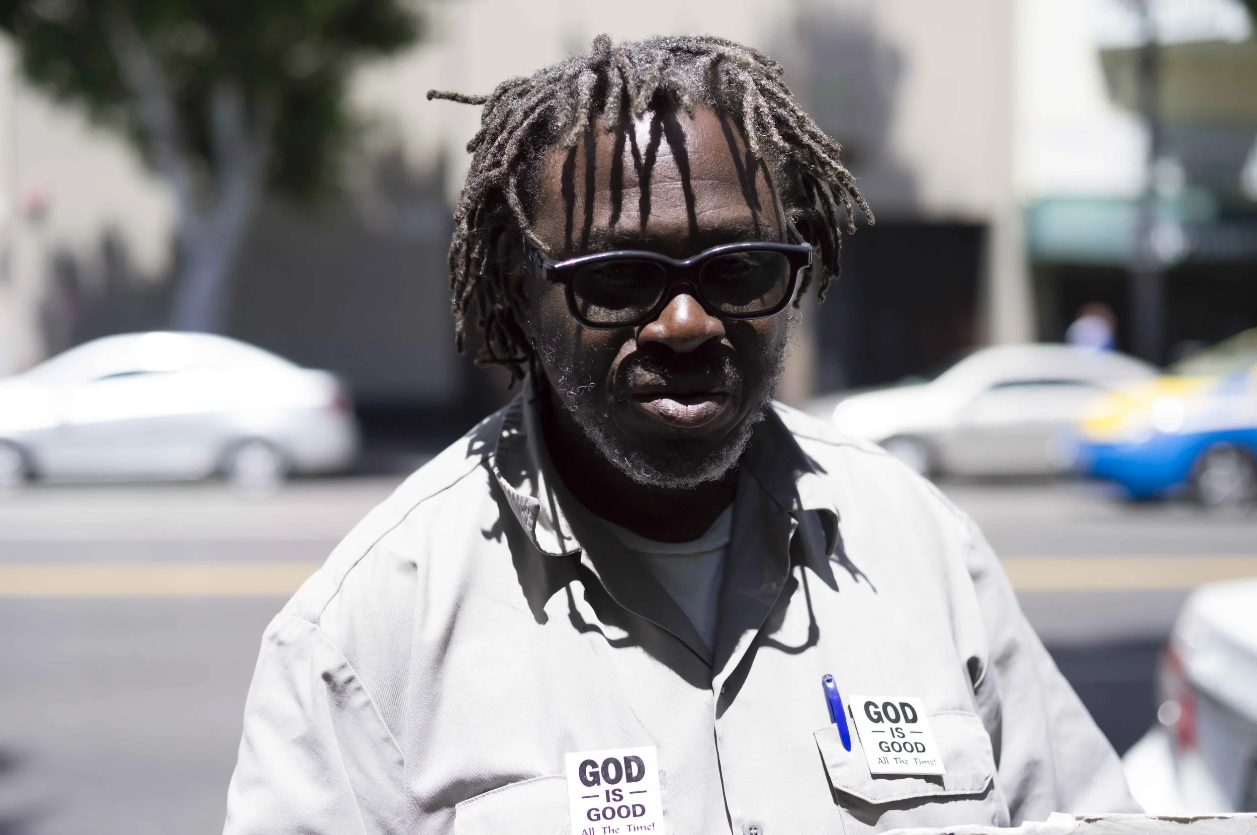 A man wearing sunglasses and a white shirt with the phrase 'GOD IS GOOD' on note cards attached to his shirt. He is outdoors in a sunny urban environment.