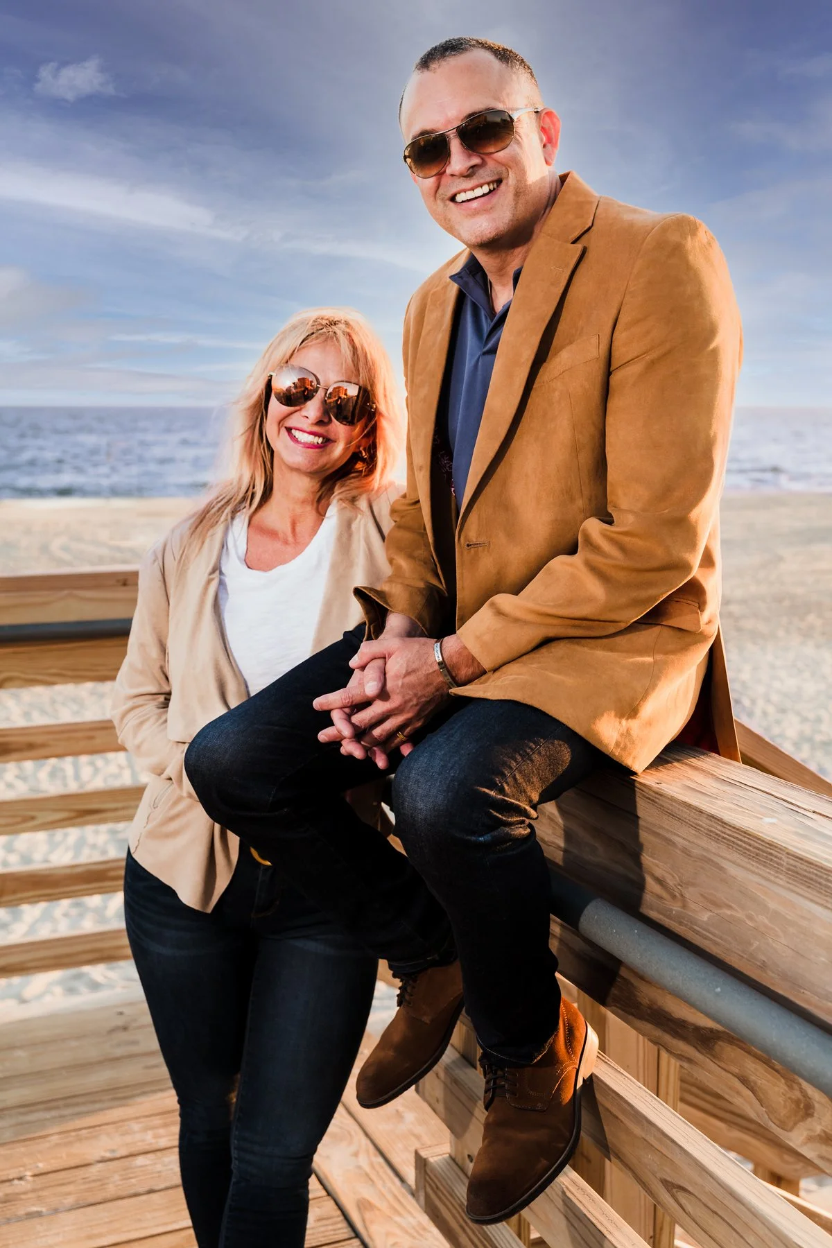 A smiling man and woman wearing sunglasses sitting on a wooden railing at the beach during sunset.