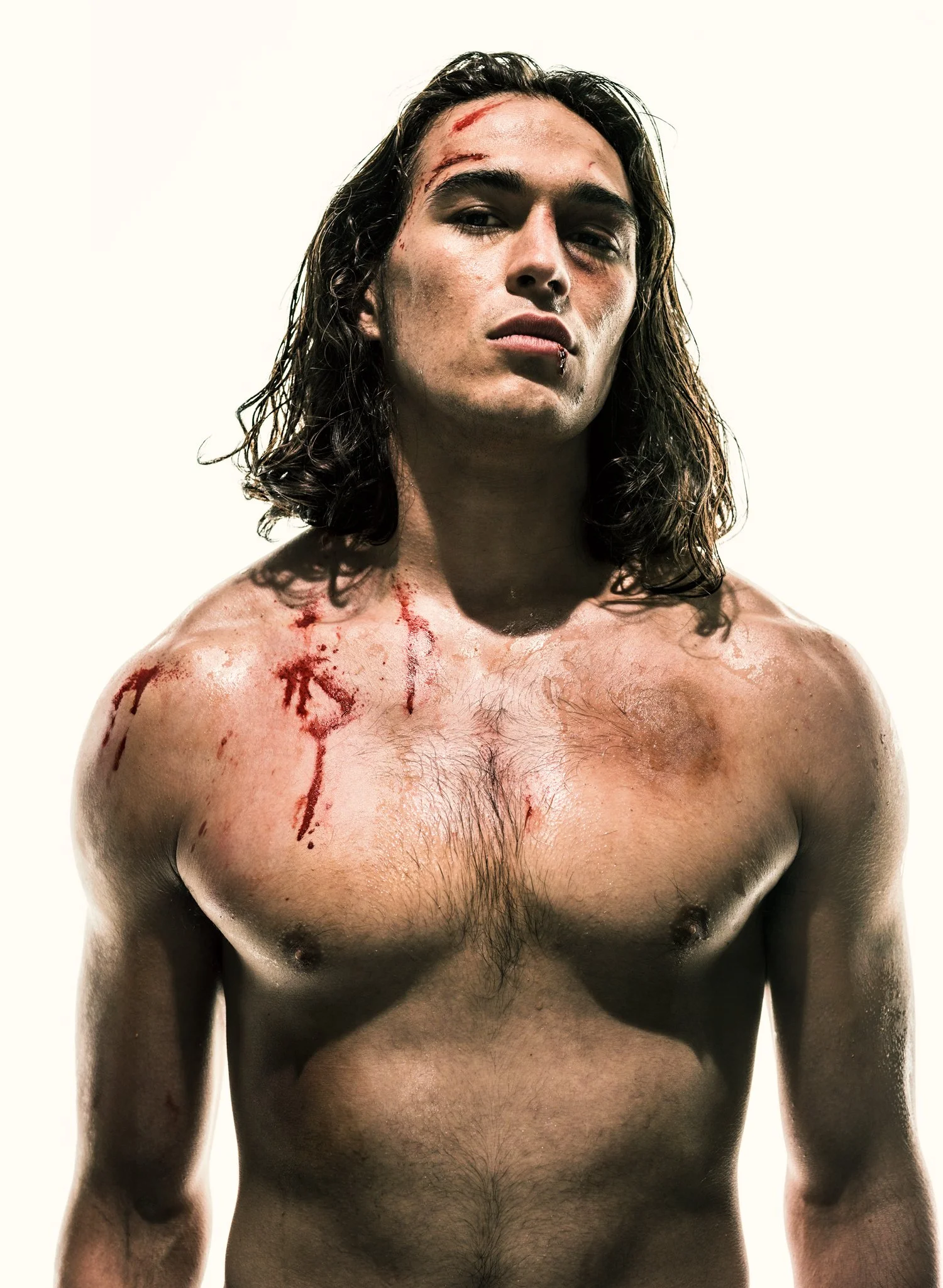 A shirtless man with long, wavy hair and injuries on his face and shoulders, some of which are bleeding, against a plain white background.