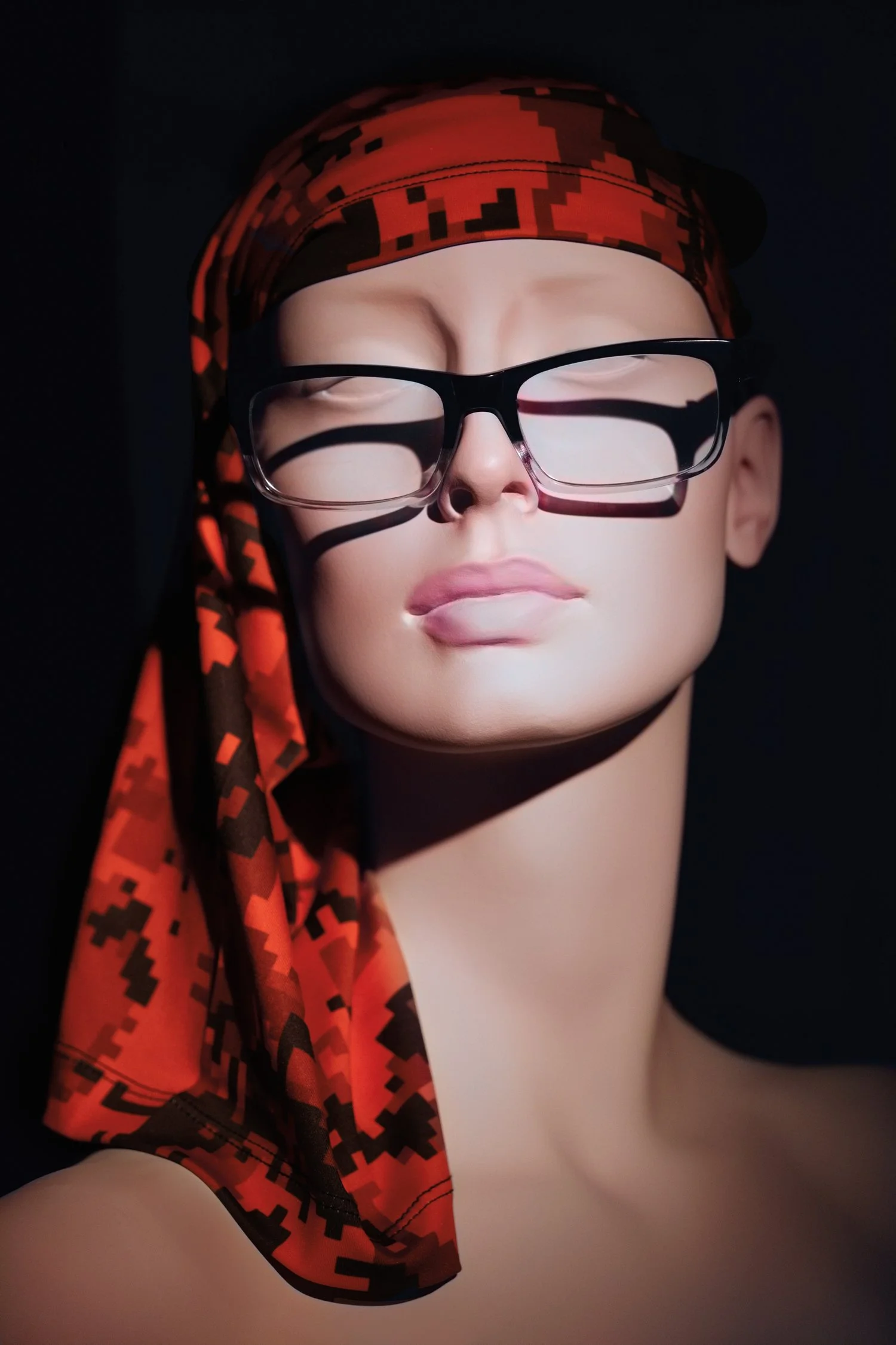 Close-up of a mannequin head wearing black-rimmed glasses, a red and black digital camouflage headscarf, and a matching scarf around the neck, with a black background.