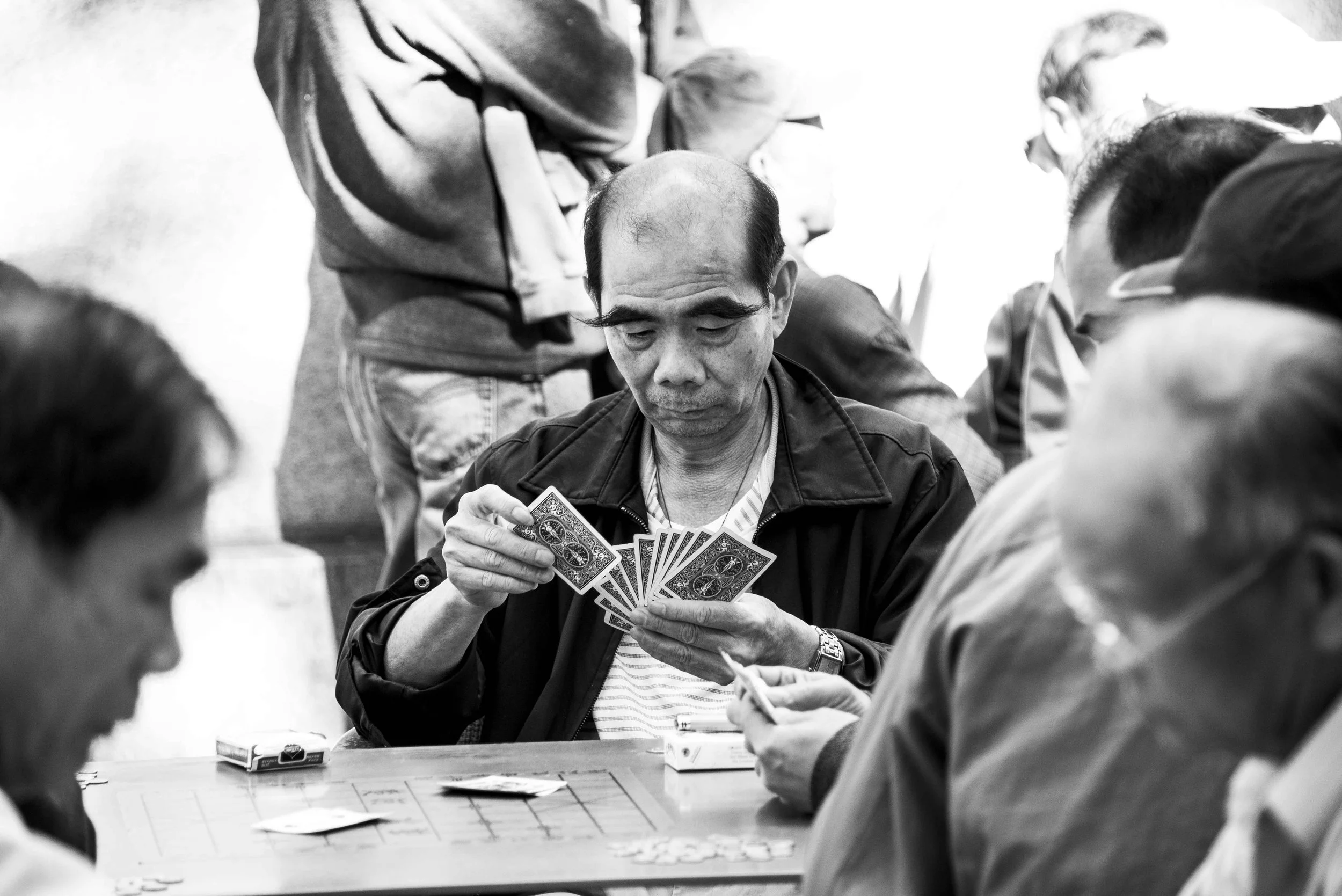 A man playing cards with a group of people around a table, holding playing cards, in a social gathering.