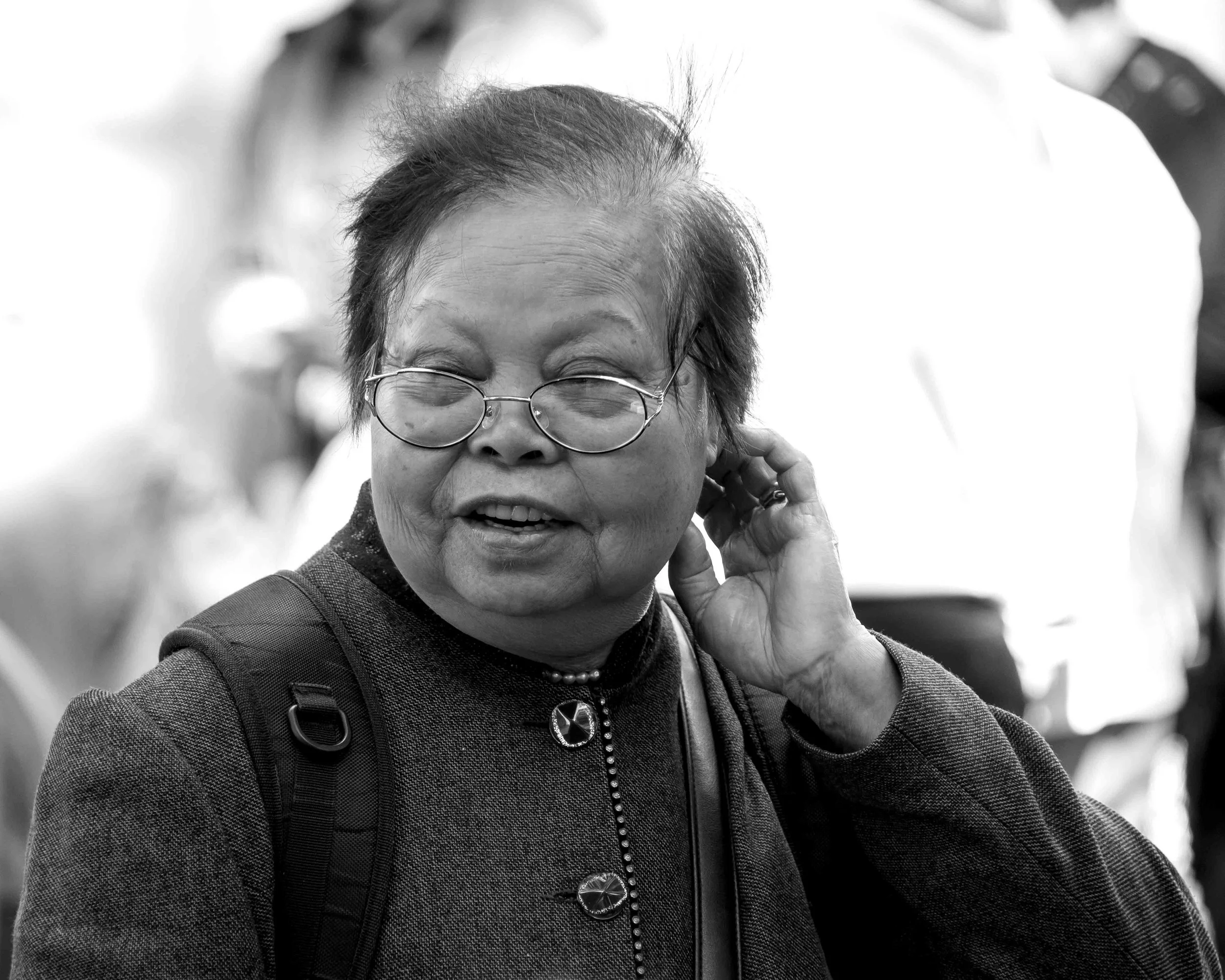 A woman with short hair, glasses, and a patterned jacket, smiling and adjusting her earring. She has a necklace with a circular pendant and is carrying a backpack.