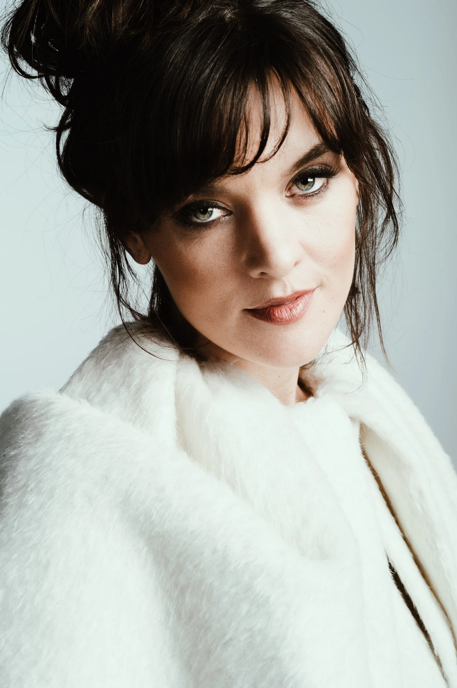 Actor Frankie Shaw with piercing green eyes, her hair in a messy up-do, depicted in a closeup portrait wearing a textured, white shawl and staring directly into the camera with head tilted to the left. Shot on a neutral, pale blue background. with di