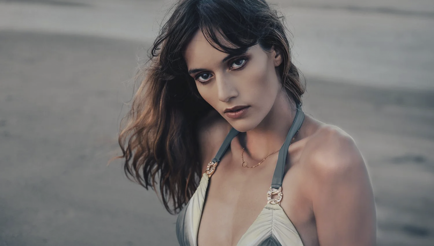 A young woman with dark wavy hair and piercing blue eyes gazes directly at the camera, standing outdoors in a natural setting with a blurred background.