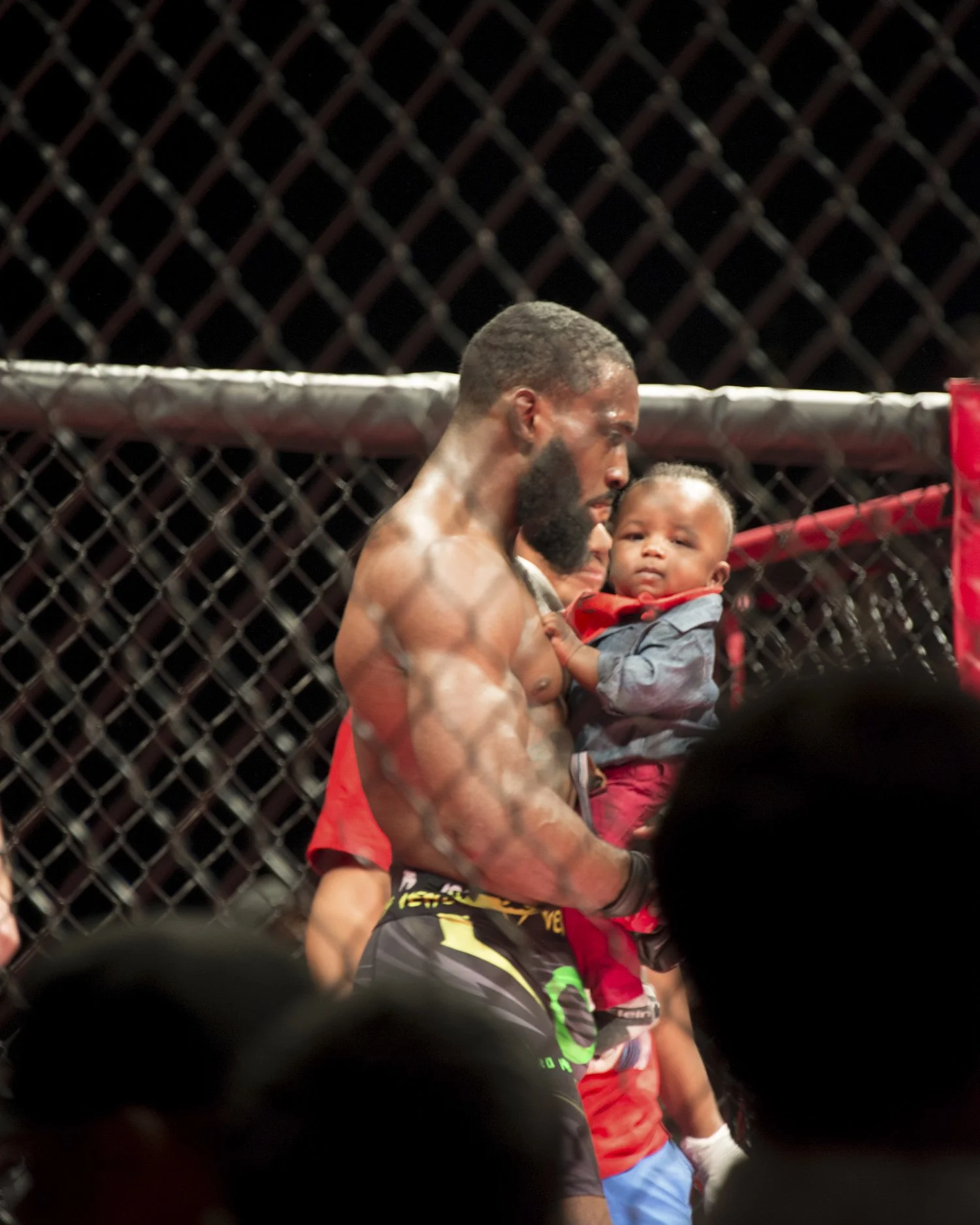 A shirtless male mixed martial arts fighter standing inside a cage, holding a young child dressed in a red shirt, against a black chain-link fence background.