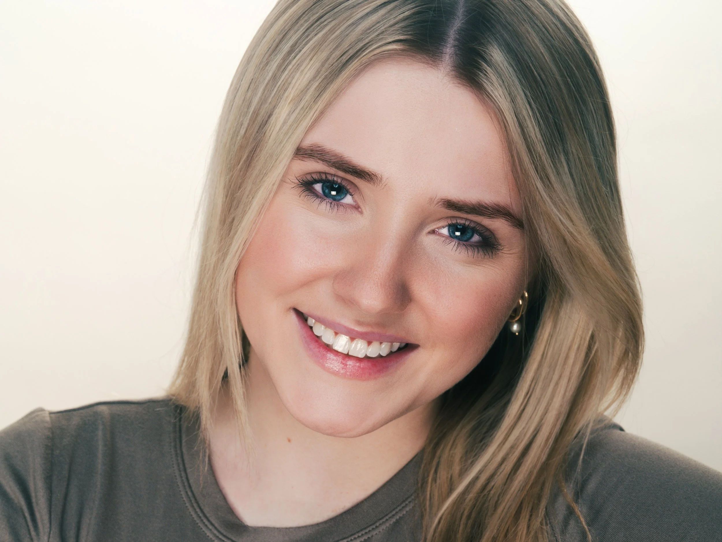 A young, smiling woman with long, straight blonde hair, sparkling blue eyes, and engaging smile. She is wearing a dark gray t-shirt against a warm-neutral background.