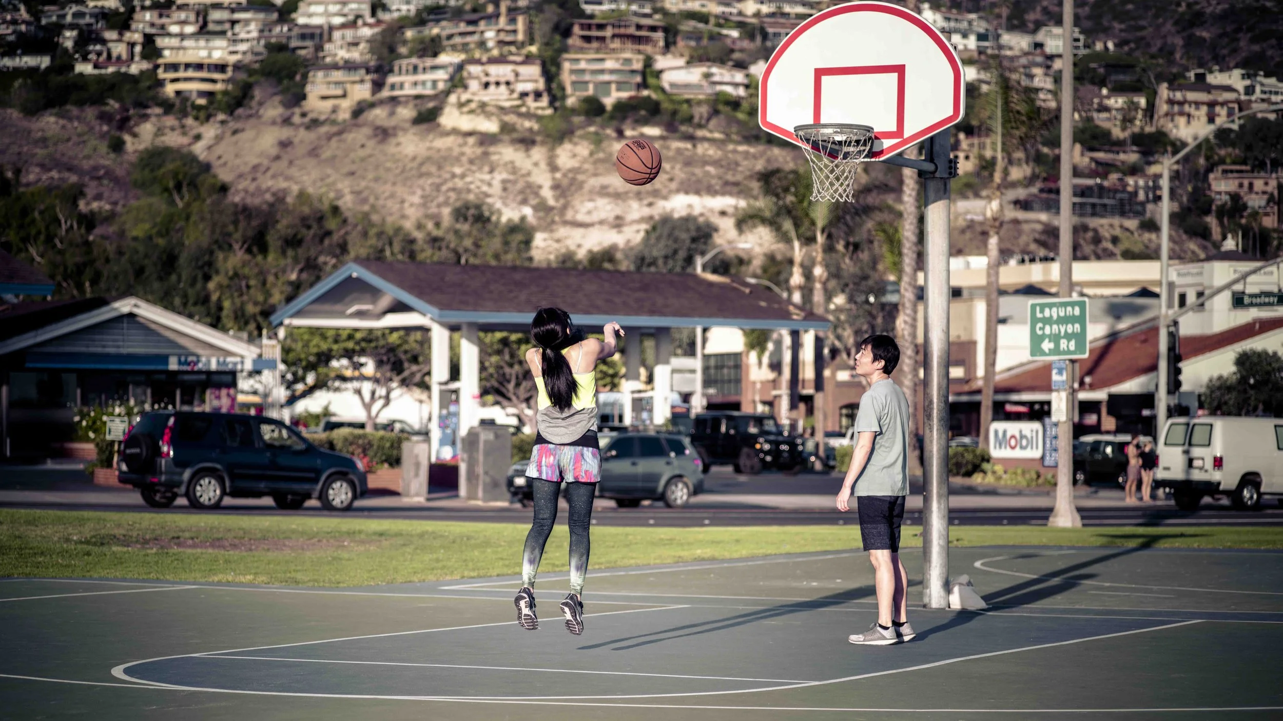 A girl is jumping to shoot a basketball on an outdoor basketball court while a boy stands nearby watching. There are parked cars, trees, and houses on a hillside in the background.