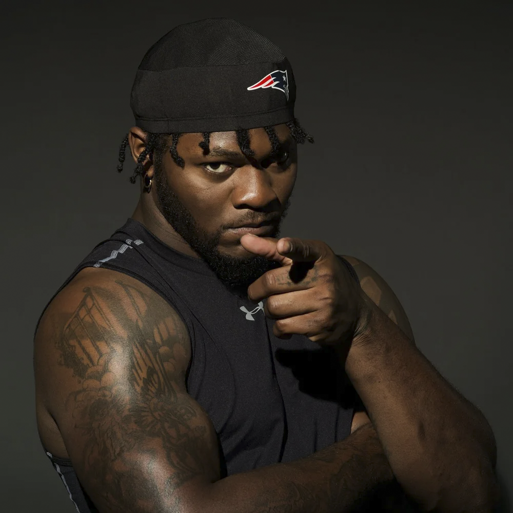 A muscular man with tattoos on his arm, wearing a black sleeveless shirt and a black cap with a sports team logo, pointing directly at the camera with an intense expression.