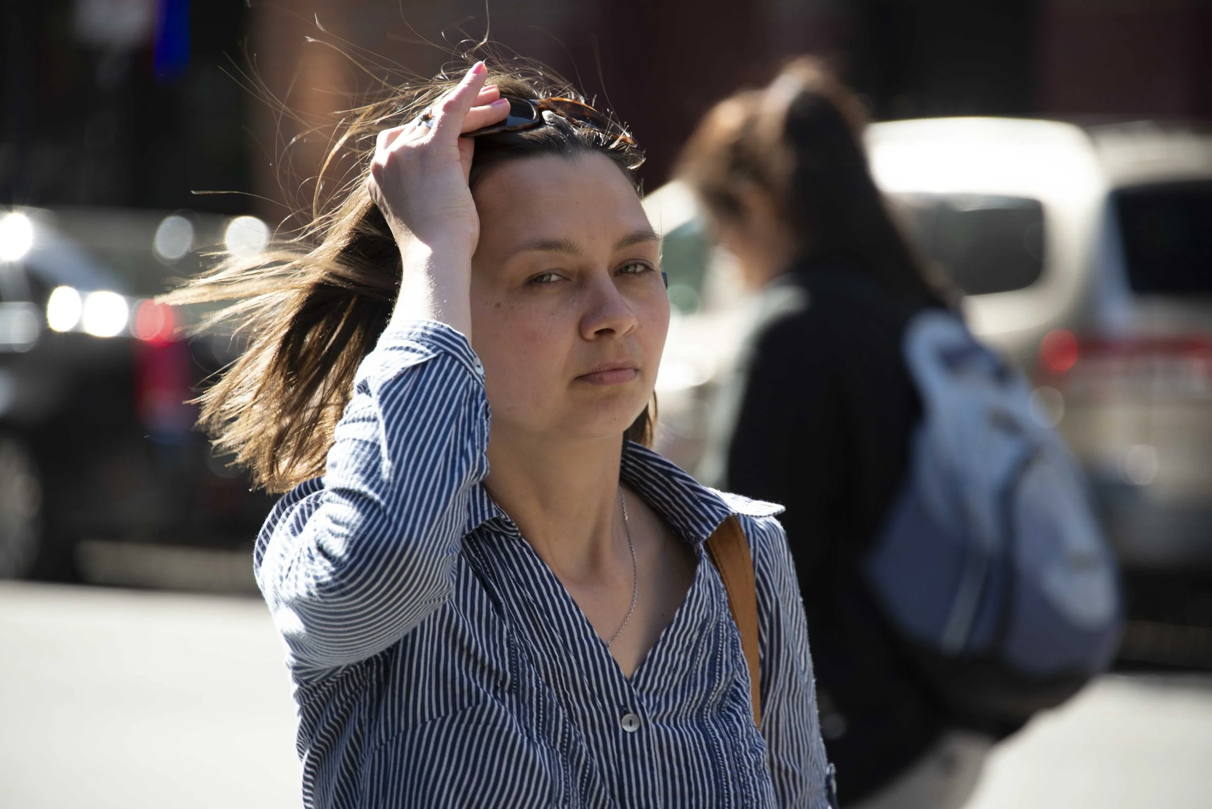 A young woman with long brown hair, wearing a striped shirt, is adjusting her sunglasses while walking outdoors on a sunny day. There are blurred cars and another person in the background.