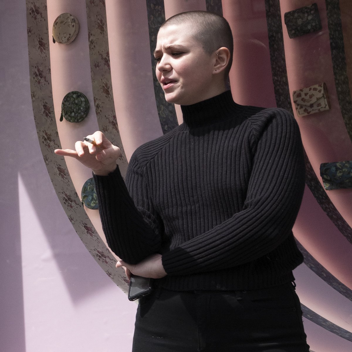 A person with a shaved head wearing a black ribbed turtleneck sweater standing against a decorative wall with floral patterned plates and small handbags. The person is holding a small object in their right hand and appears to be speaking or explainin