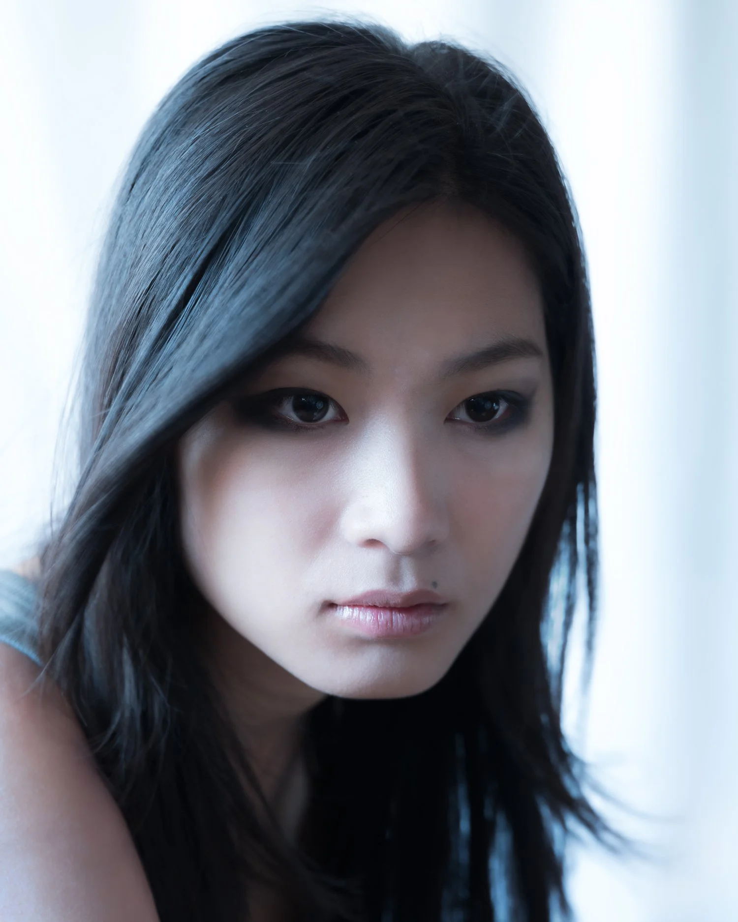 Closeup portrait of young Asian woman in high-contrast, bluish natural light against a white fabric backdrop. She has pale skin with long, black hair and wears heavy eye makeup, looking away from the camera in a moment of introspection.