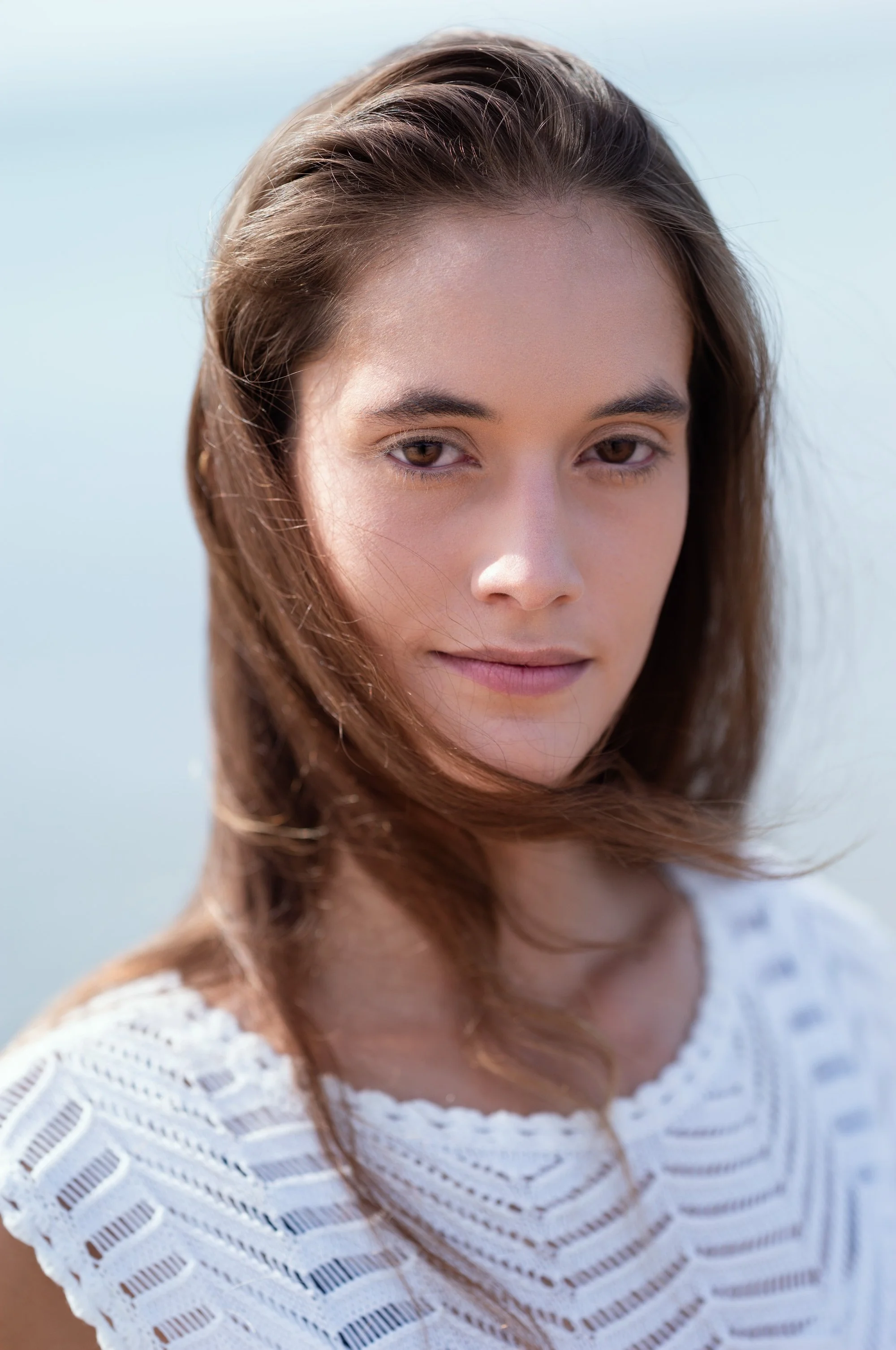 Close-up of a young woman with brown, wavy hair, wearing a white, cut-out knit top, outdoors in natural light.