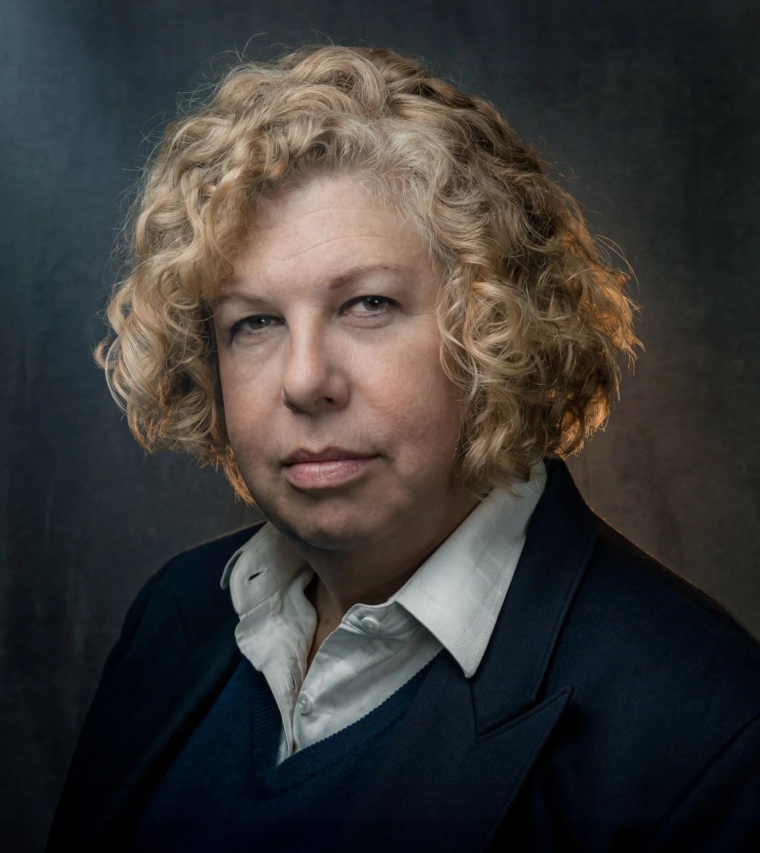 Portrait of a trans woman with curly, blonde hair, looking intently at the camera, wearing a dark blazer with a blue v-neck sweater over a white, collared shirt against a blue-gray background.