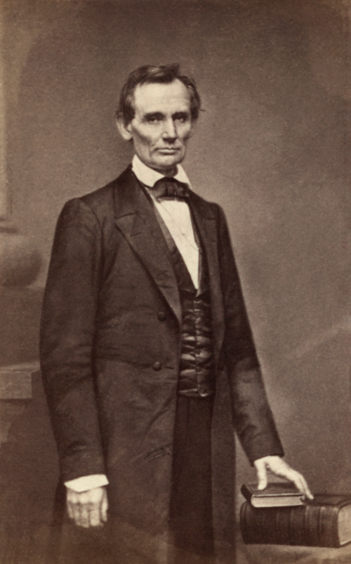 Portrait of Abraham Lincoln, standing, wearing a black overcoat, posing with his left hand resting on a stack of books.