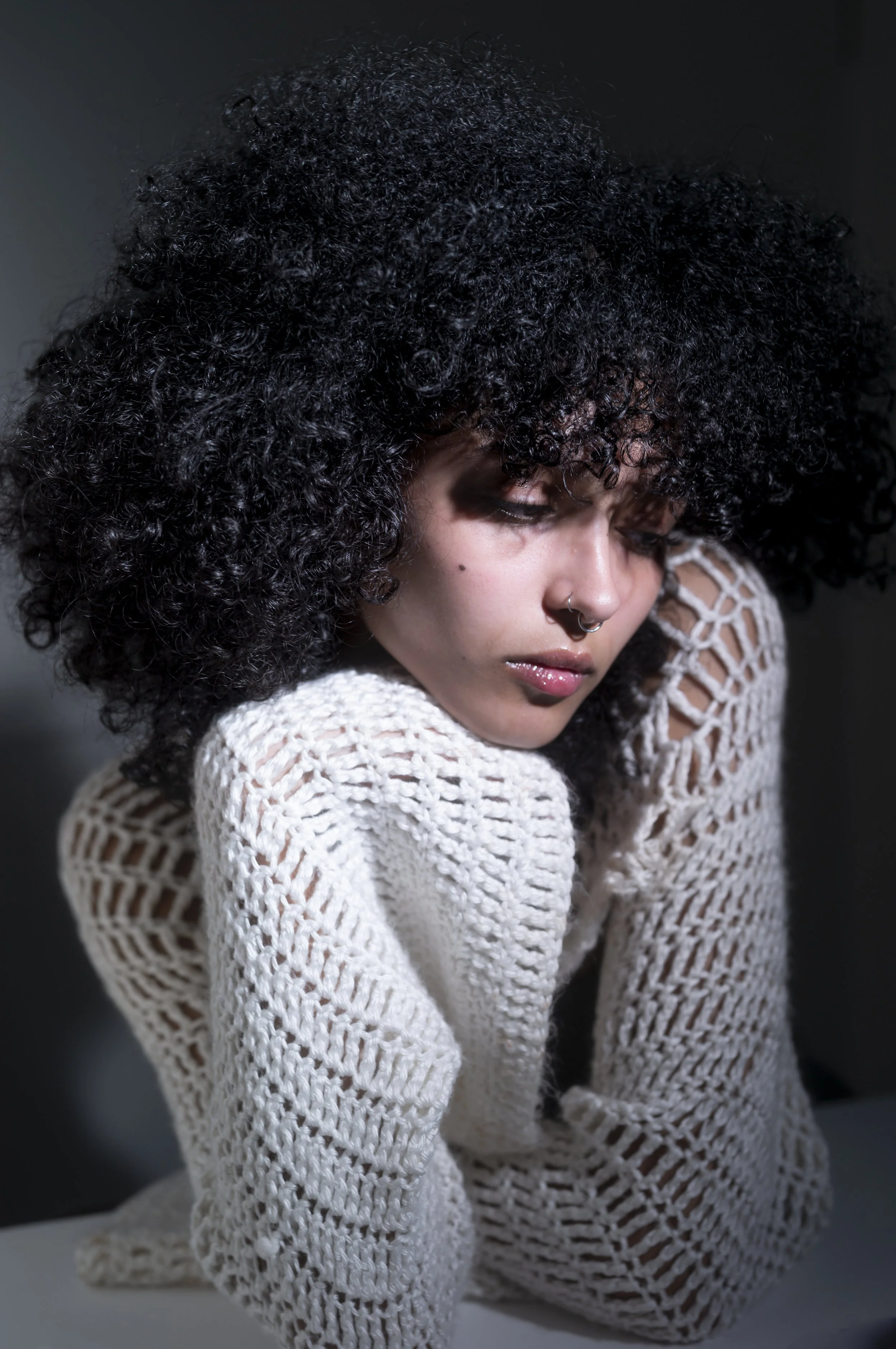 A woman with curly black hair, wearing a white crocheted sweater, resting her head on her arm, looking down with a thoughtful expression.