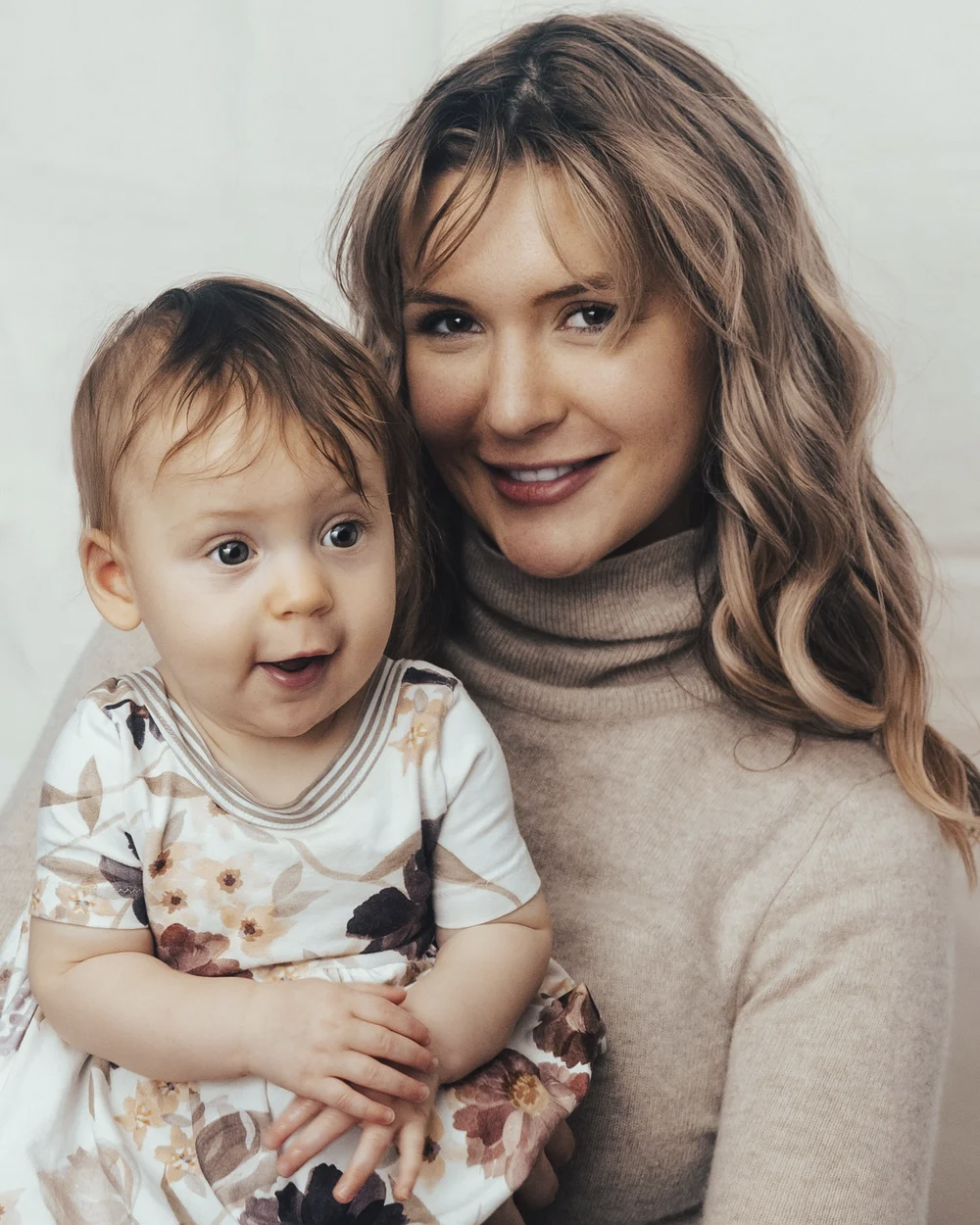A woman with long wavy hair holding a smiling infant girl with short hair and blue eyes, both looking happy and close together.