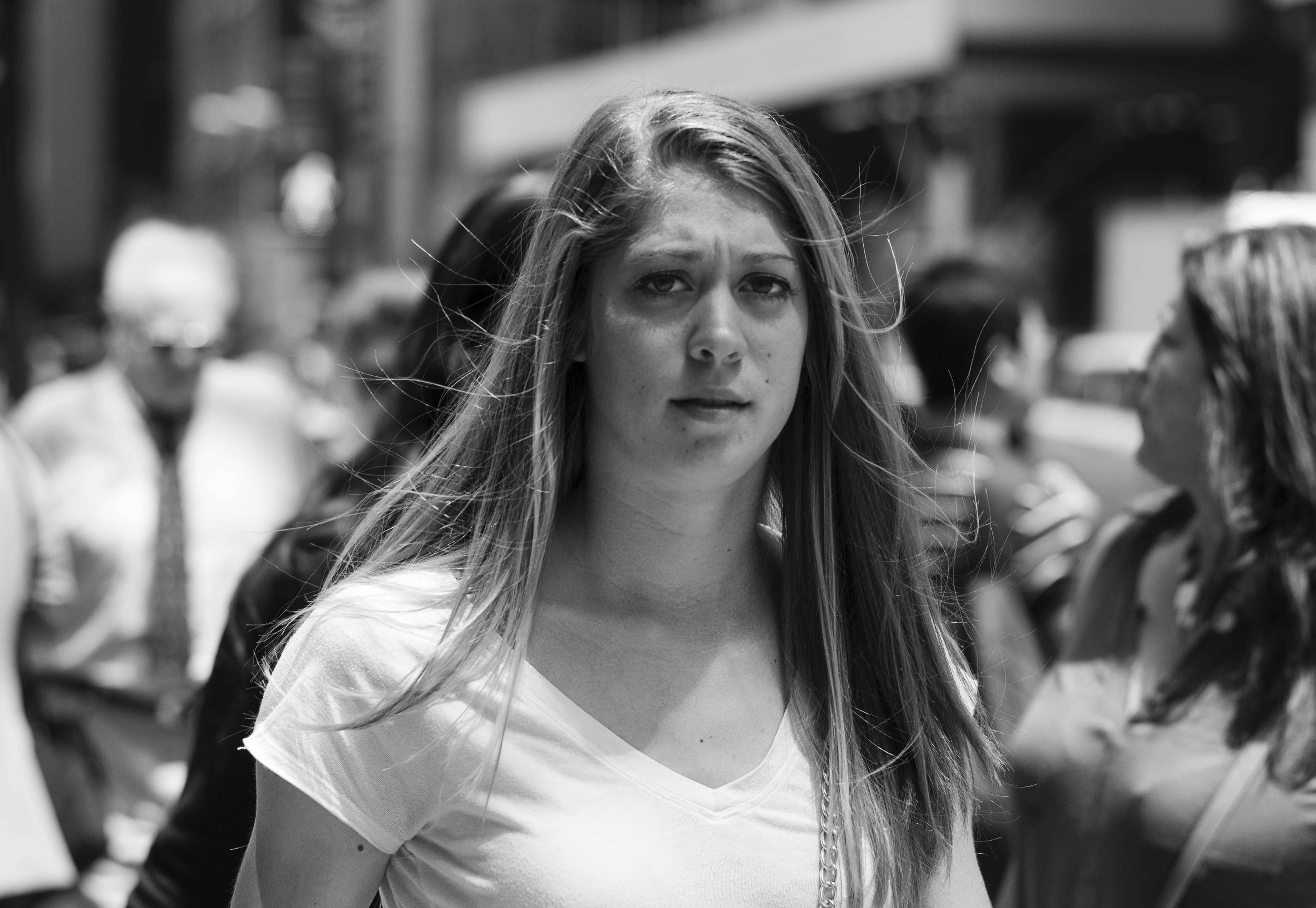 Black-and-white photo of a young woman with long hair looking at the camera in a busy outdoor setting, with blurred people and objects in the background.
