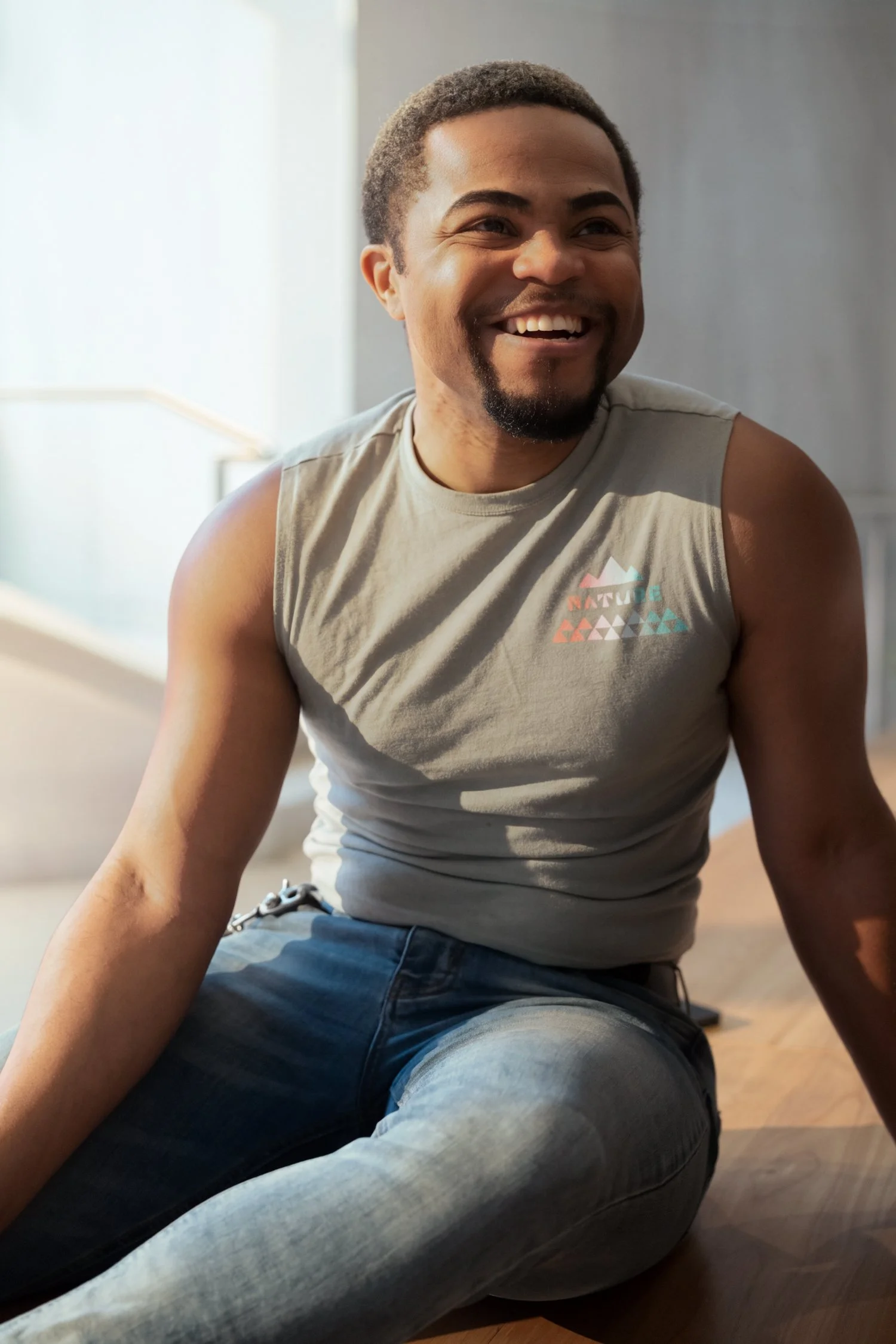 Smiling man sitting on the floor in a well-lit room, wearing a gray sleeveless athletic shirt with colorful triangle logo and blue jeans.