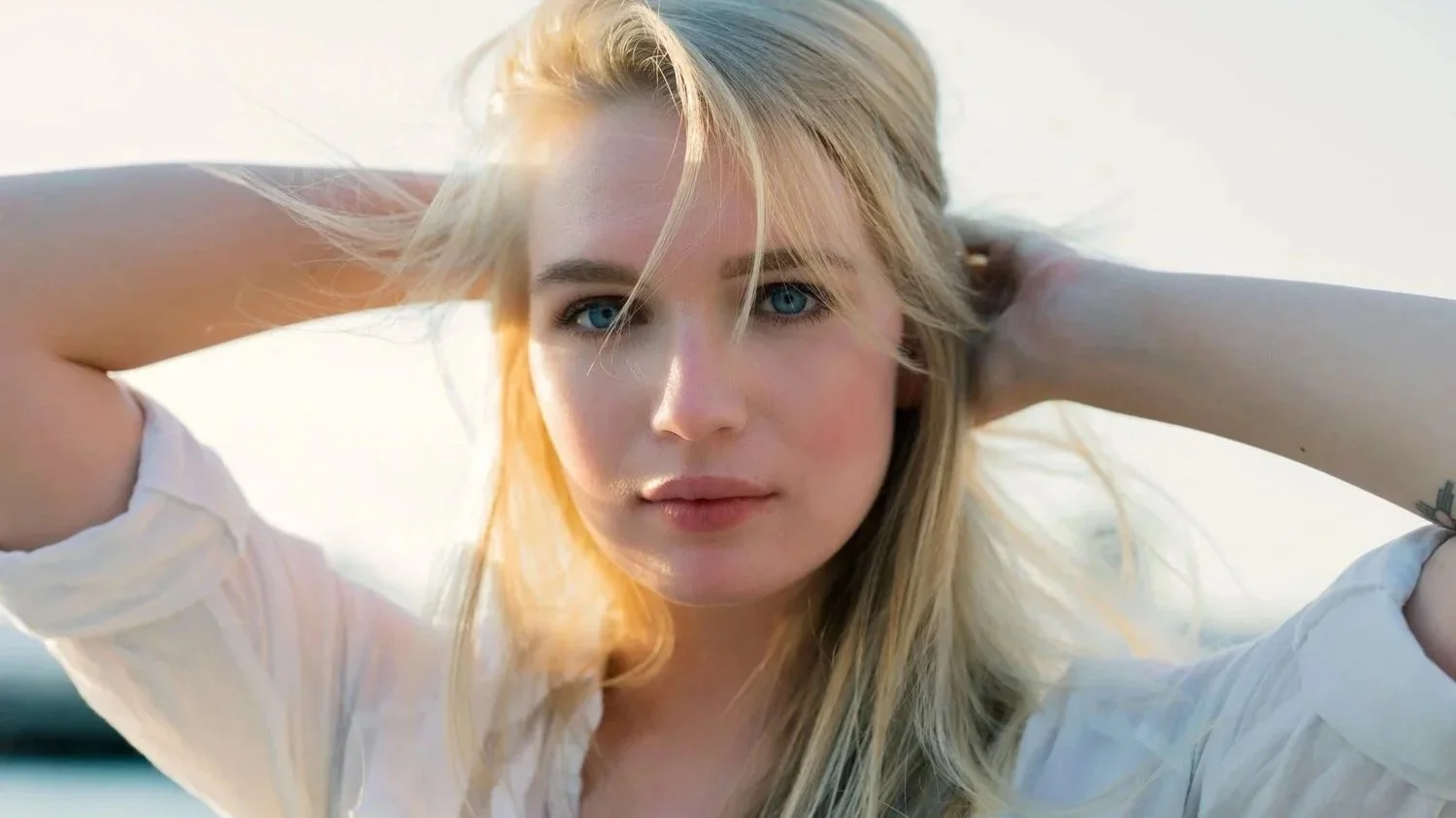 A young woman with long blonde hair and blue eyes looking directly at the camera, with arms raised behind her head and sunlight illuminating her face.