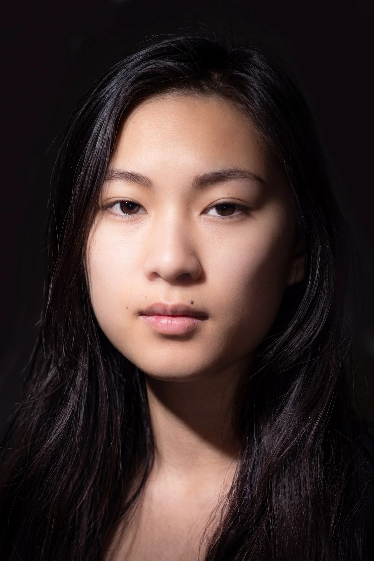 Studio portrait of young, expressionless woman with long, dark hair and pale skin against a black background with soft, directional light and deep shadows.