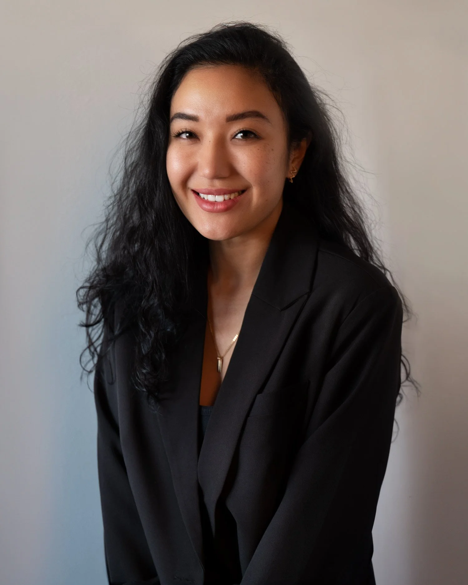 Headshot of a woman with long, black, curly hair, wearing a black blazer and gold jewelry, smiling against a plain wall background.