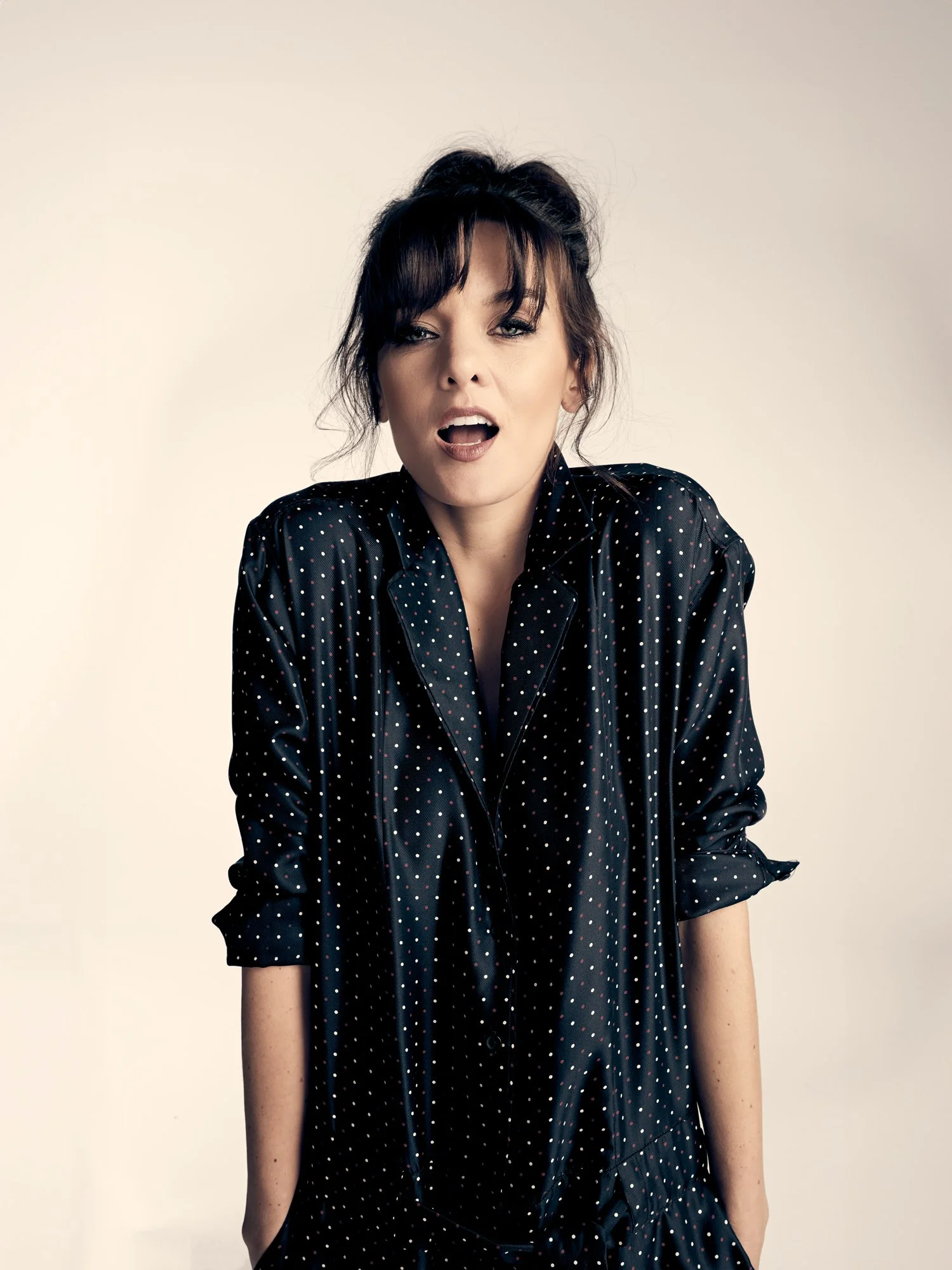 Actor Frankie Shaw with her dark hair pinned up, emotes with expressive body-language dressed in a dark blue jumper with a white and pink dot pattern, shot against a warm, neutral background.