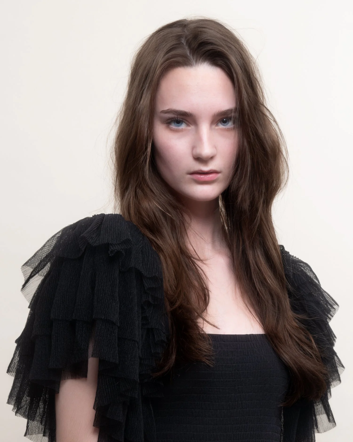 A young woman with long brown hair and blue eyes, wearing a black top with ruffled tulle sleeves, standing against a plain off-white background.