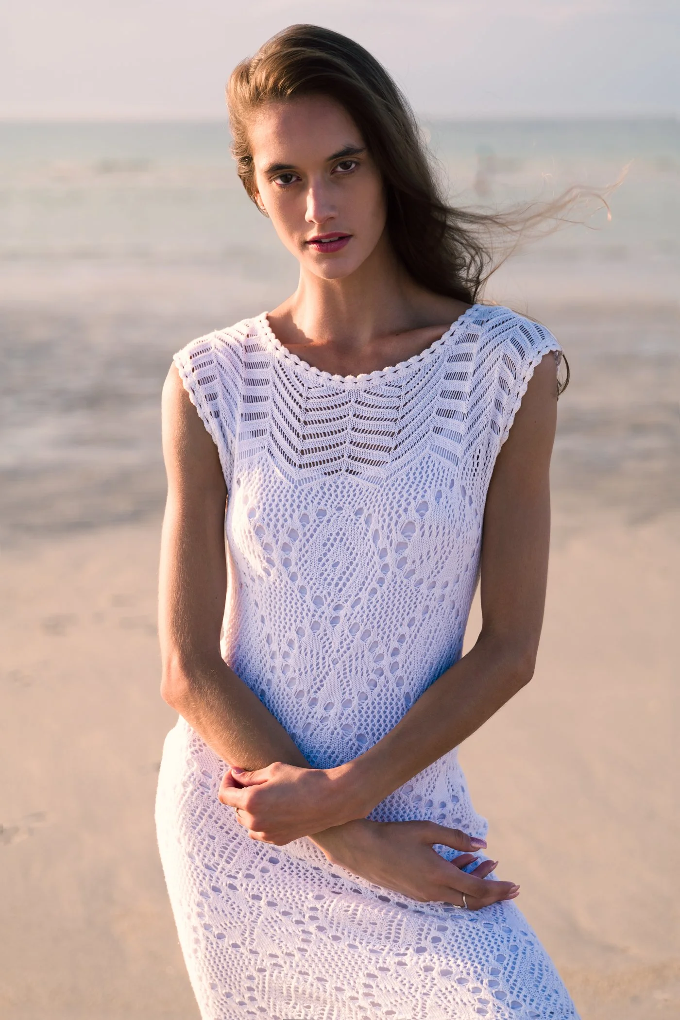 A woman in a white crocheted dress standing on a beach with the ocean in the background, wind blowing her hair, looking at the camera.
