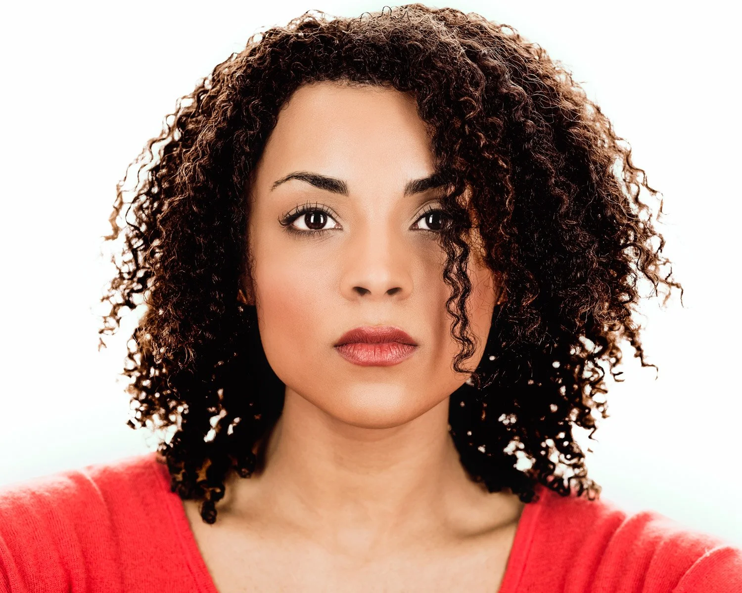 A Black woman with dark curly hair, wearing a red top, looking directly into the camera with a neutral expression against a white background.