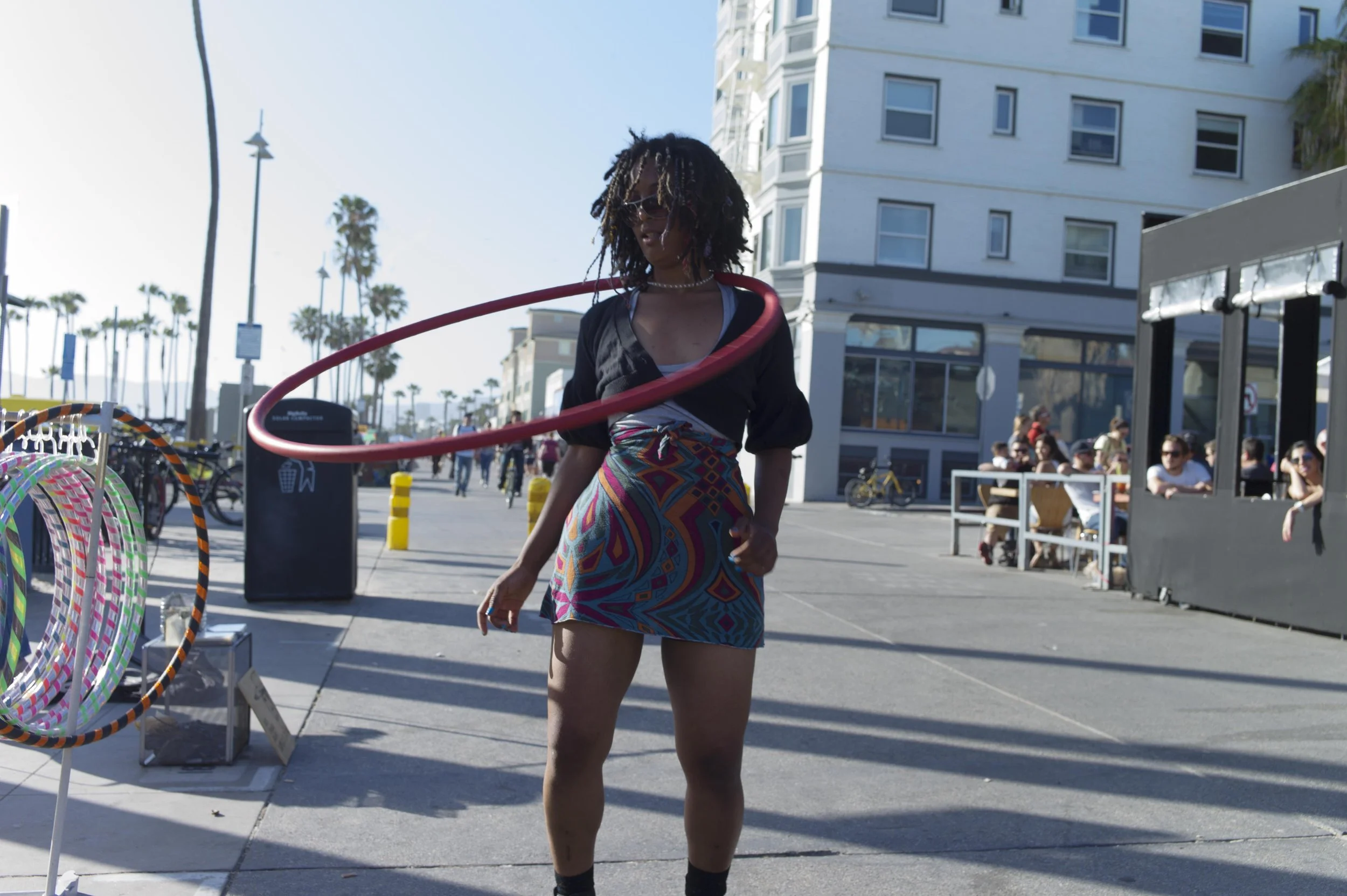 A woman with dreadlocks, sunglasses, a black blouse, and a colorful skirt is walking on a busy outdoor street with palm trees, modern buildings, and people sitting on benches in the background.