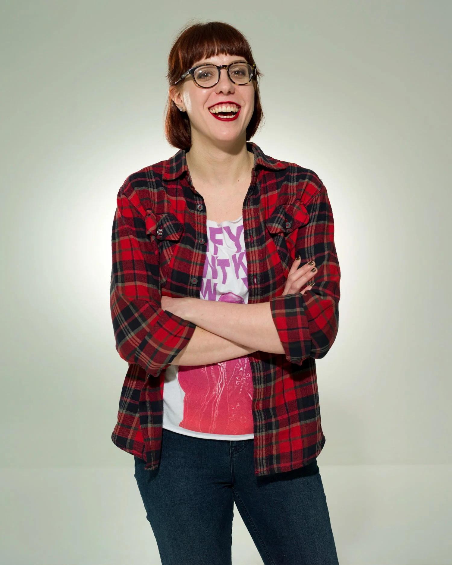 A woman with short brown hair, glasses, and red lipstick, smiling with arms crossed, wearing a red and black plaid shirt over a white graphic T-shirt, and dark jeans, standing against a plain light-colored background.
