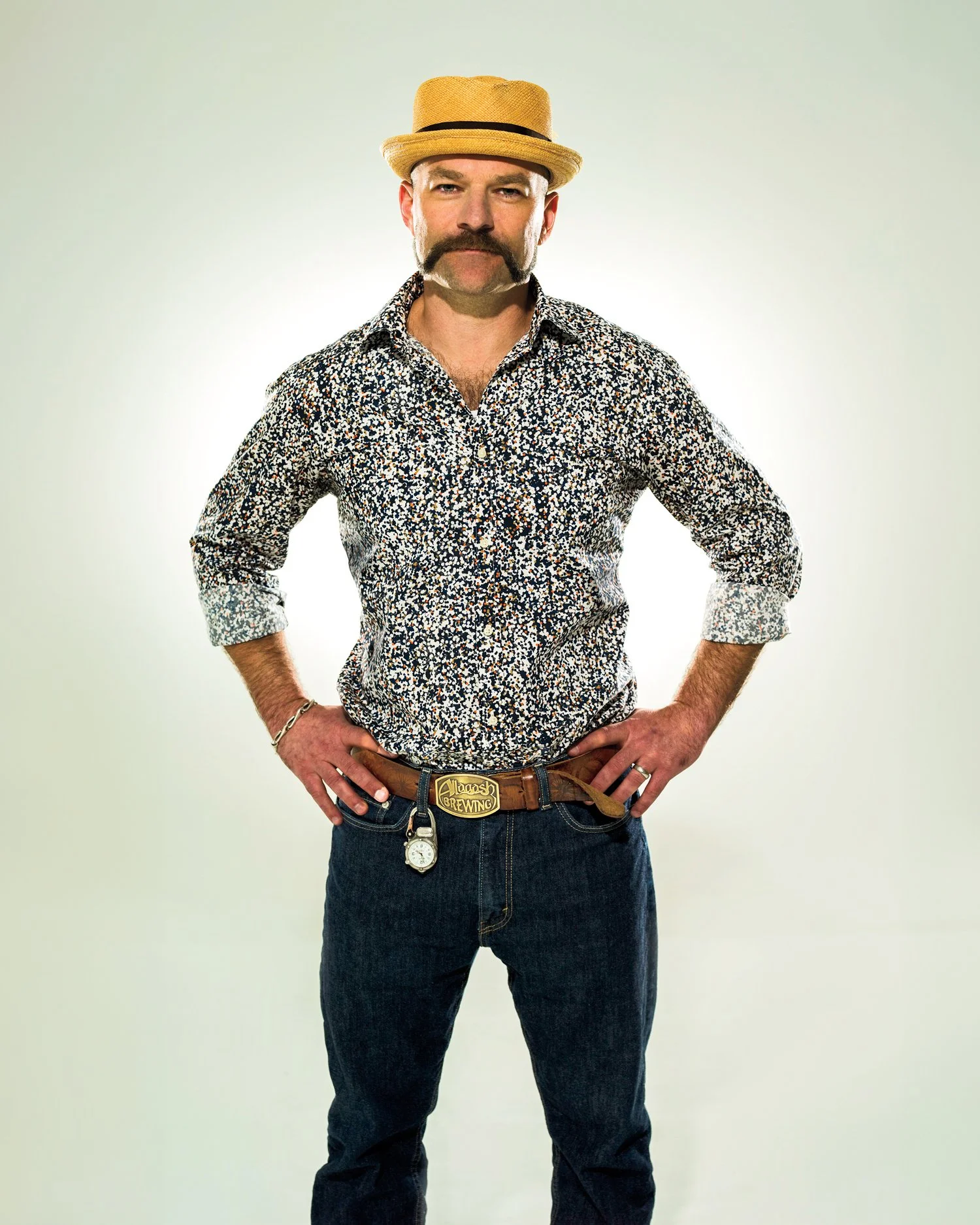 Man with a mustache and beard wearing a straw hat, floral shirt, jeans, with a belt that says 'Alagash Brewing', and a pocket watch hanging from his belt.