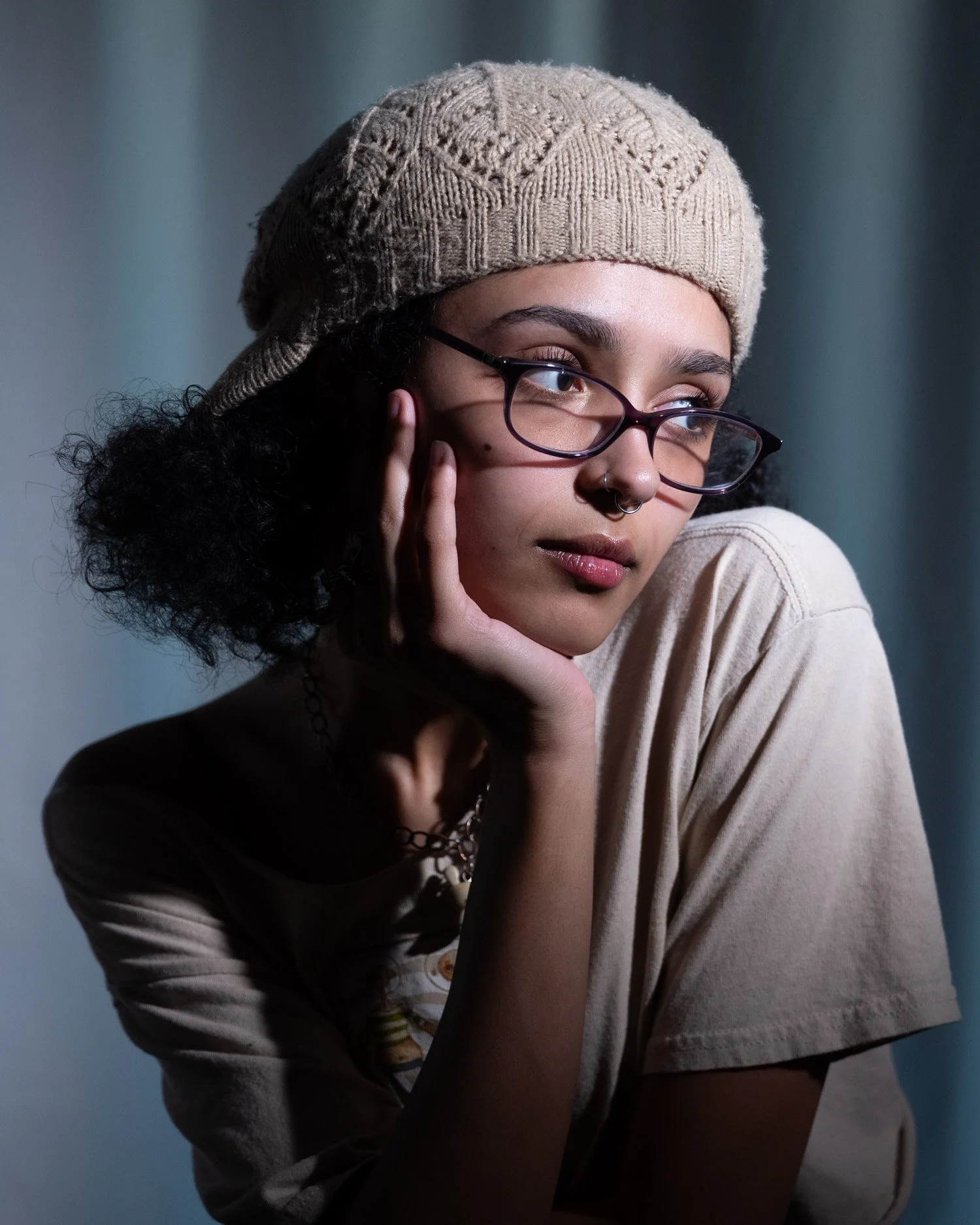 A young woman with glasses and a septum piercing resting her chin on her hand, wearing a beige beanie and a beige t-shirt.