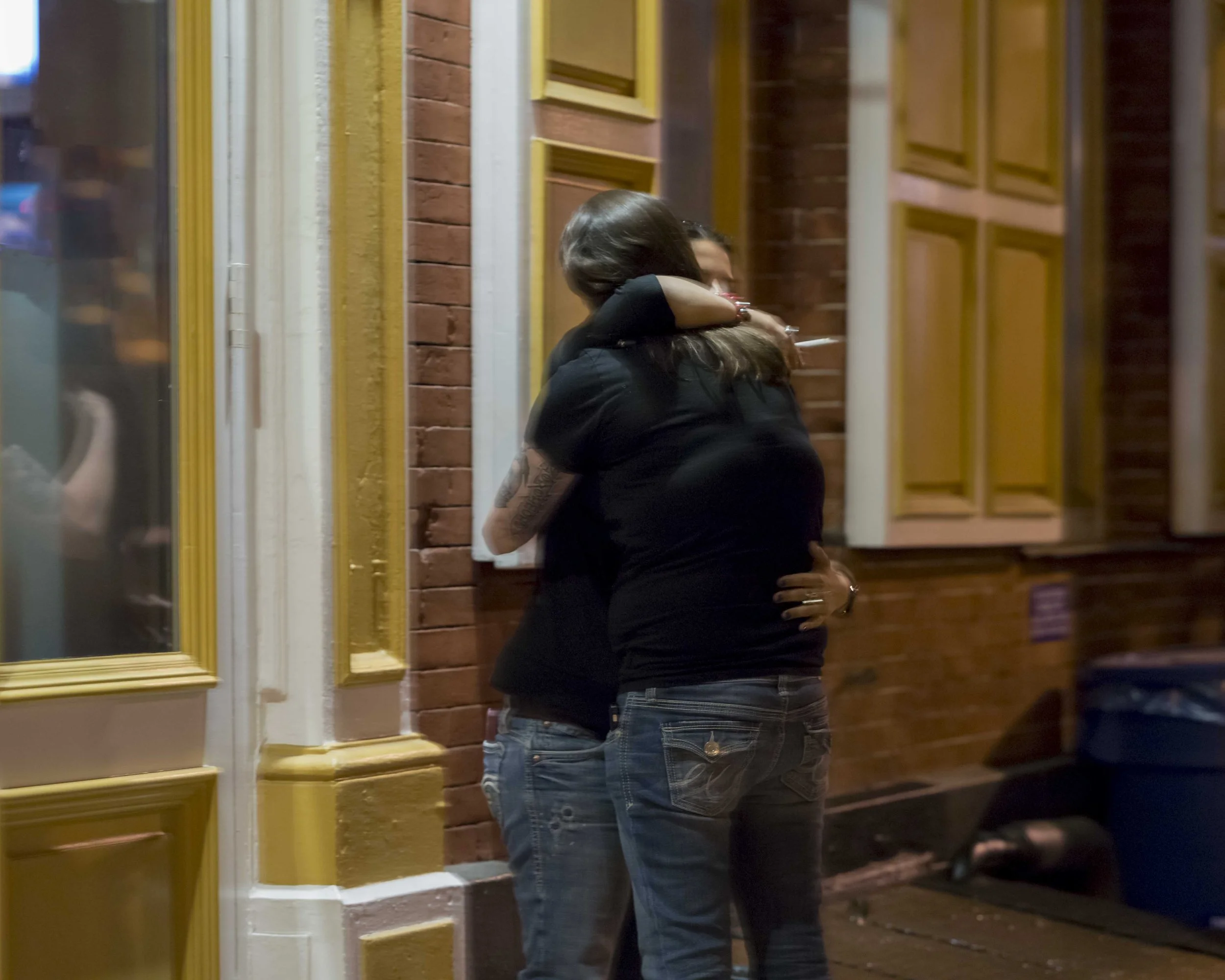 A couple embracing outside a restaurant or cafe at night, with brick and yellow-styled window frames in the background.