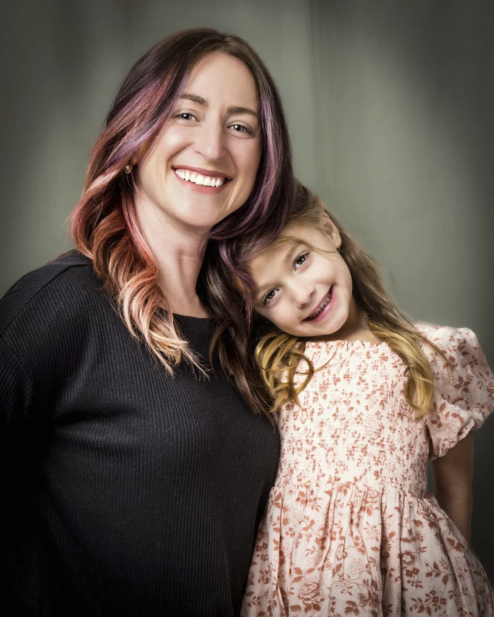 A woman and a young girl standing close together, smiling, with the woman’s head leaning on the girl’s head against a neutral background.