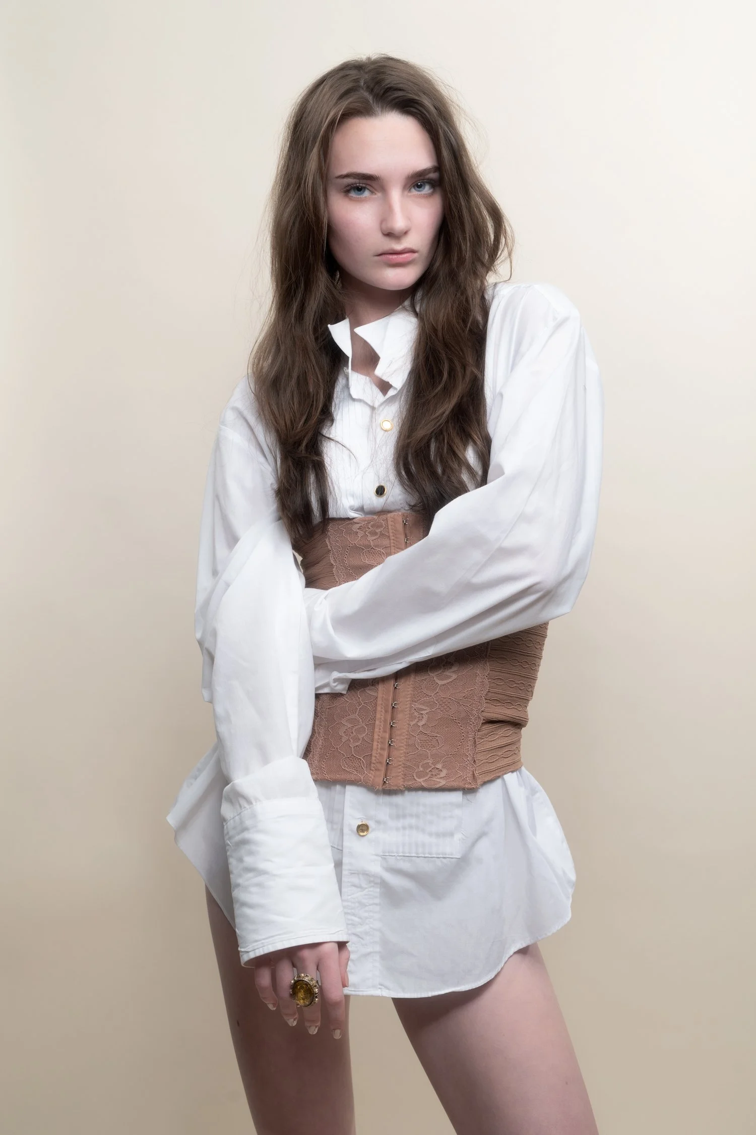 Young woman with long wavy brown hair wearing a white oversized shirt, a tan corset, and minimal makeup, standing against a plain beige background, looking directly at the camera.
