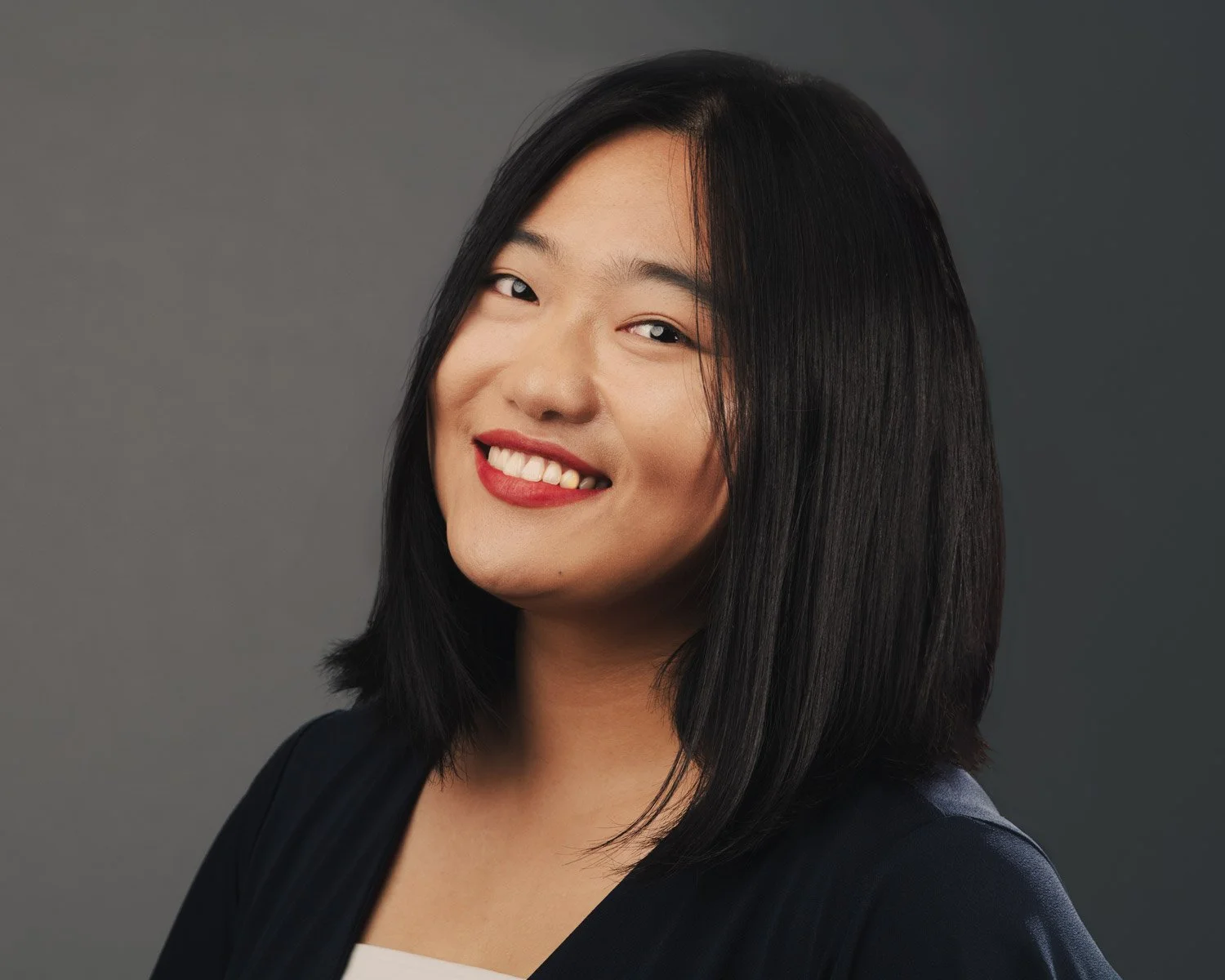 Portrait of an smiling woman with shoulder-length black hair and red lipstick, wearing a dark blazer over a white top, against a gray background.