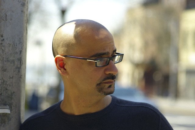 A man with glasses, a shaved head, and a goatee looking to the side outdoors on a sunny day.