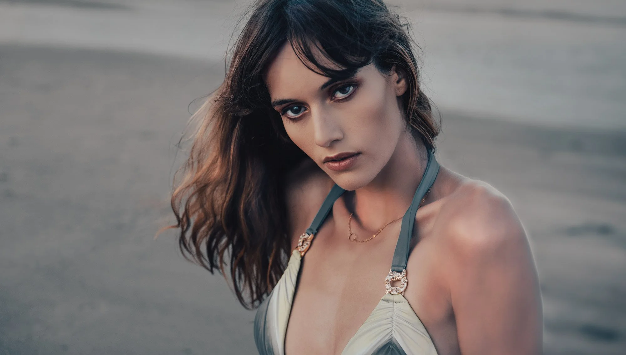A young woman with dark wavy hair and piercing blue eyes gazes directly at the camera, standing outdoors in a natural setting with a blurred background.
