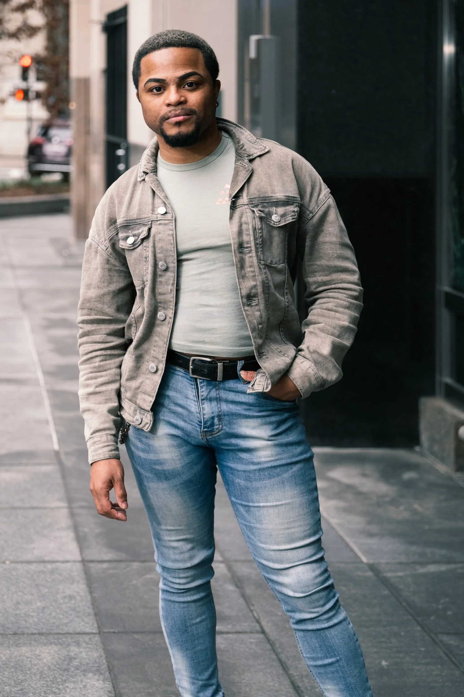 A Black man with short curly hair and a beard, wearing a tan denim jacket, light blue jeans, standing in an urban outdoor setting.