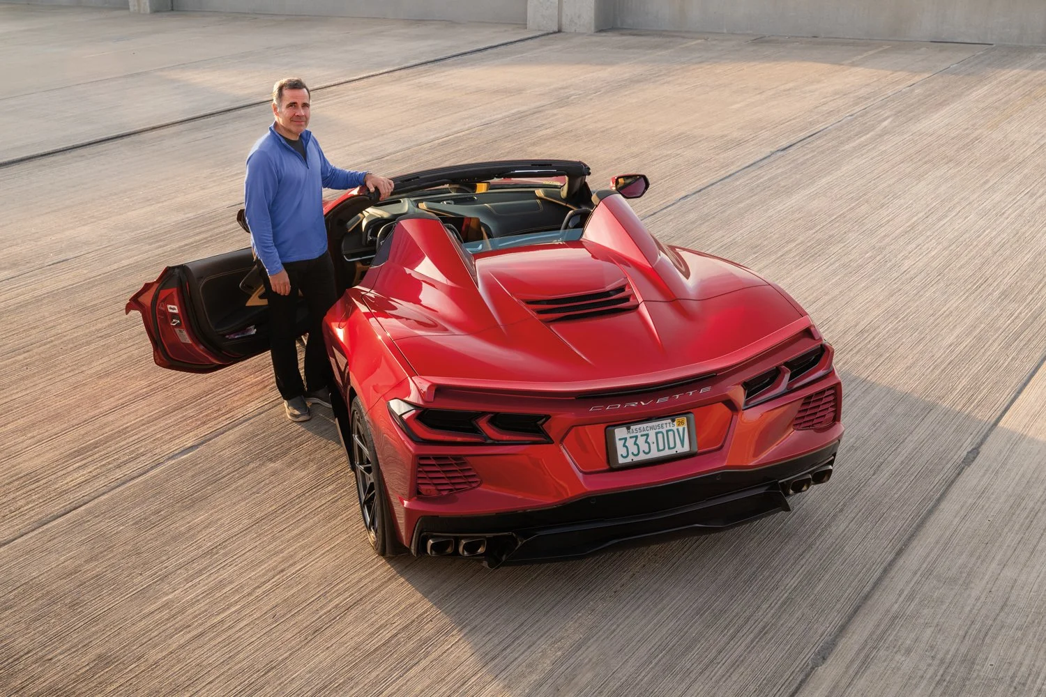 Man in a blue fleece posing next to his red Corvette Stingray on a textured concrete surface, seen from a high angle.