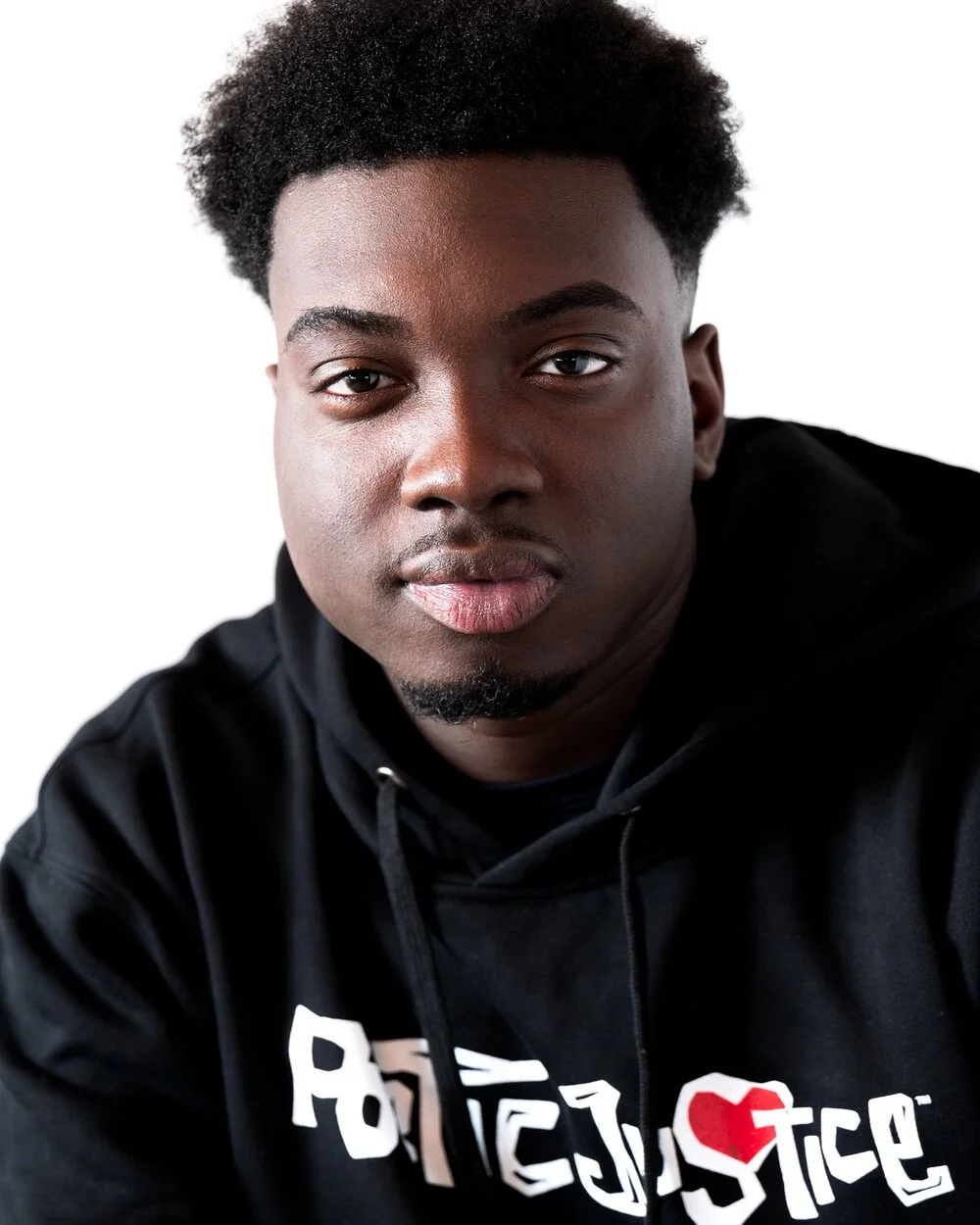 A young man with dark skin and curly hair, wearing a black hoodie with white and red text, posing against a white background.