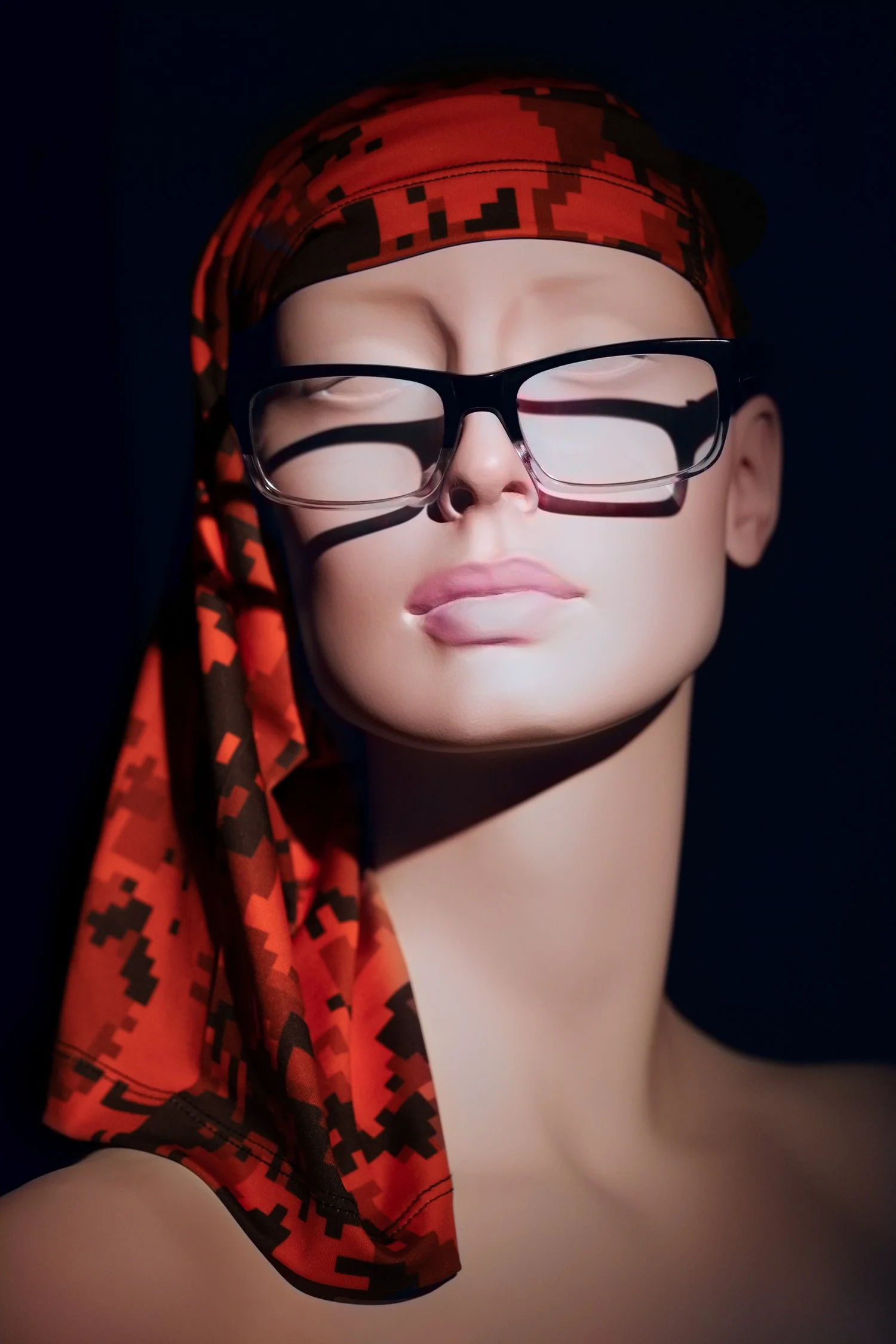 Studio mannequin portrait with orange and black camp-pattern headscarf and riding glasses, shot on a black background with hard, directional light.