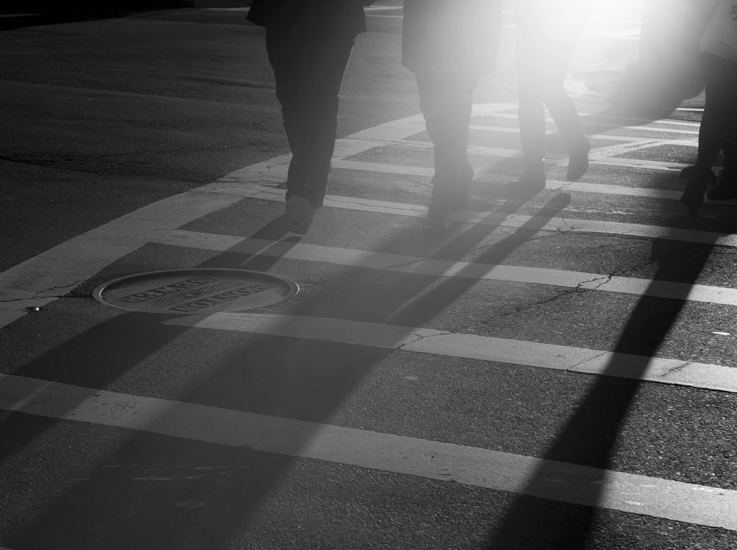 People walking across a zebra crosswalk in bright sunlight, casting shadows on the pavement.