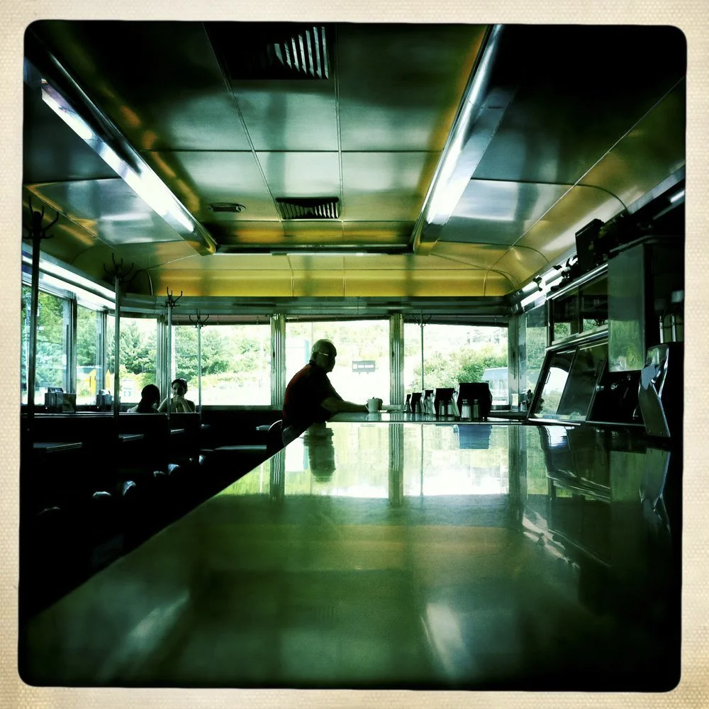 Interior of a diner or cafe with a reflective counter, large windows, and a few seated customers, with a person sitting at the counter.