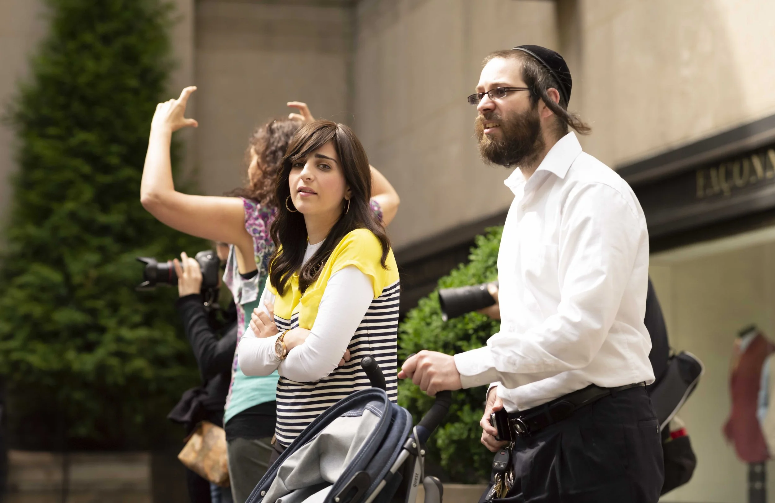 A group of people standing outdoors with greenery in the background, including a woman in a yellow and striped top and a man with a beard and glasses wearing a white shirt and a kippah, each with a stroller.