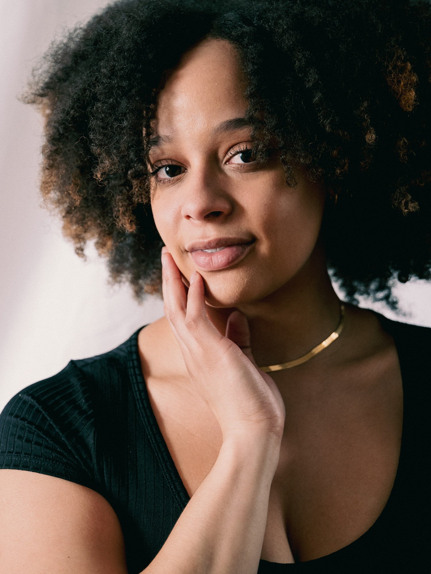 Young Black woman with dreads rests hand on her chin as she looks into the camera.
