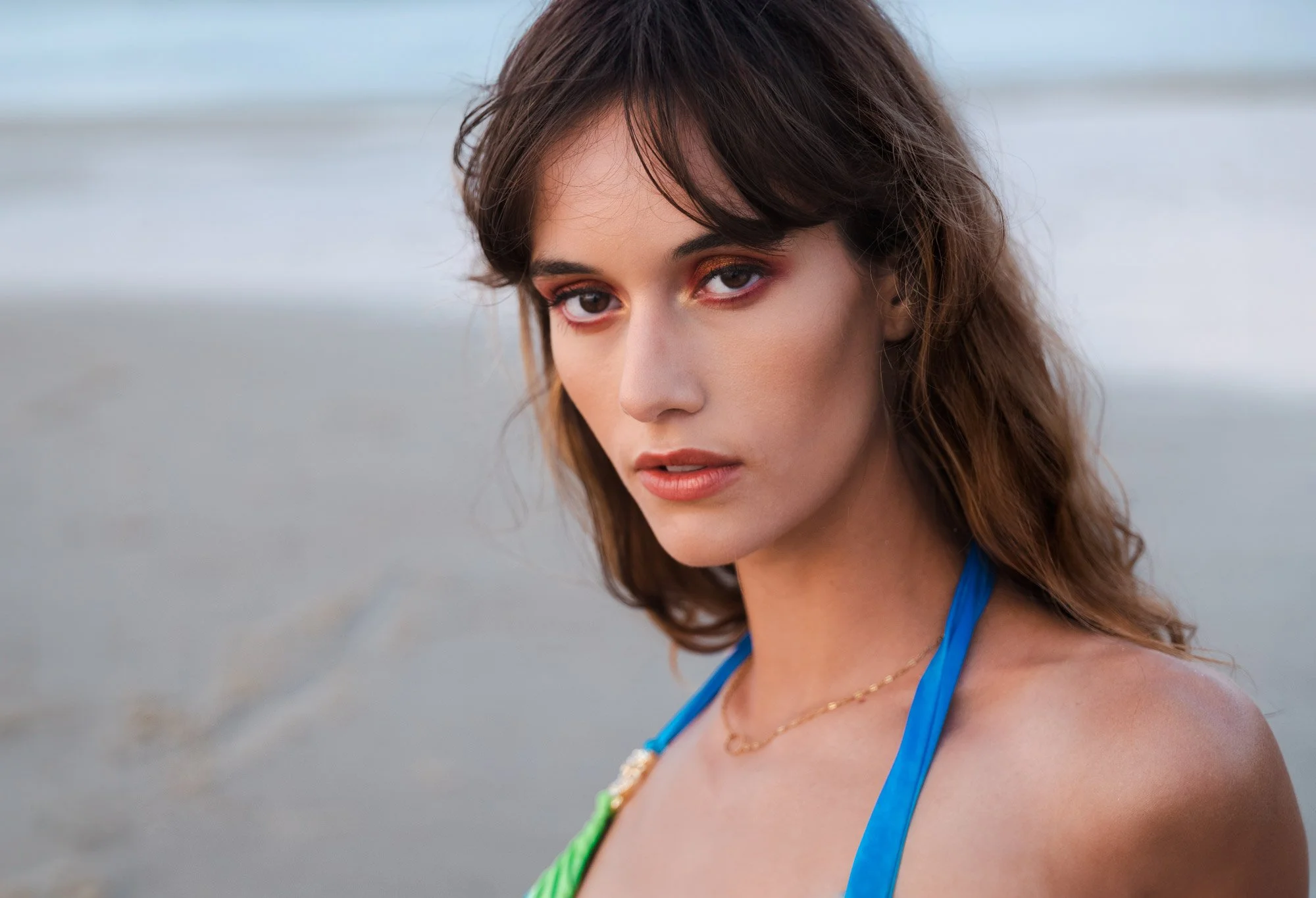 A young woman with wavy brown hair and brown eyes looking at the camera, wearing blue athletic wear with a green and yellow strap, standing outdoors near a beach or shoreline.