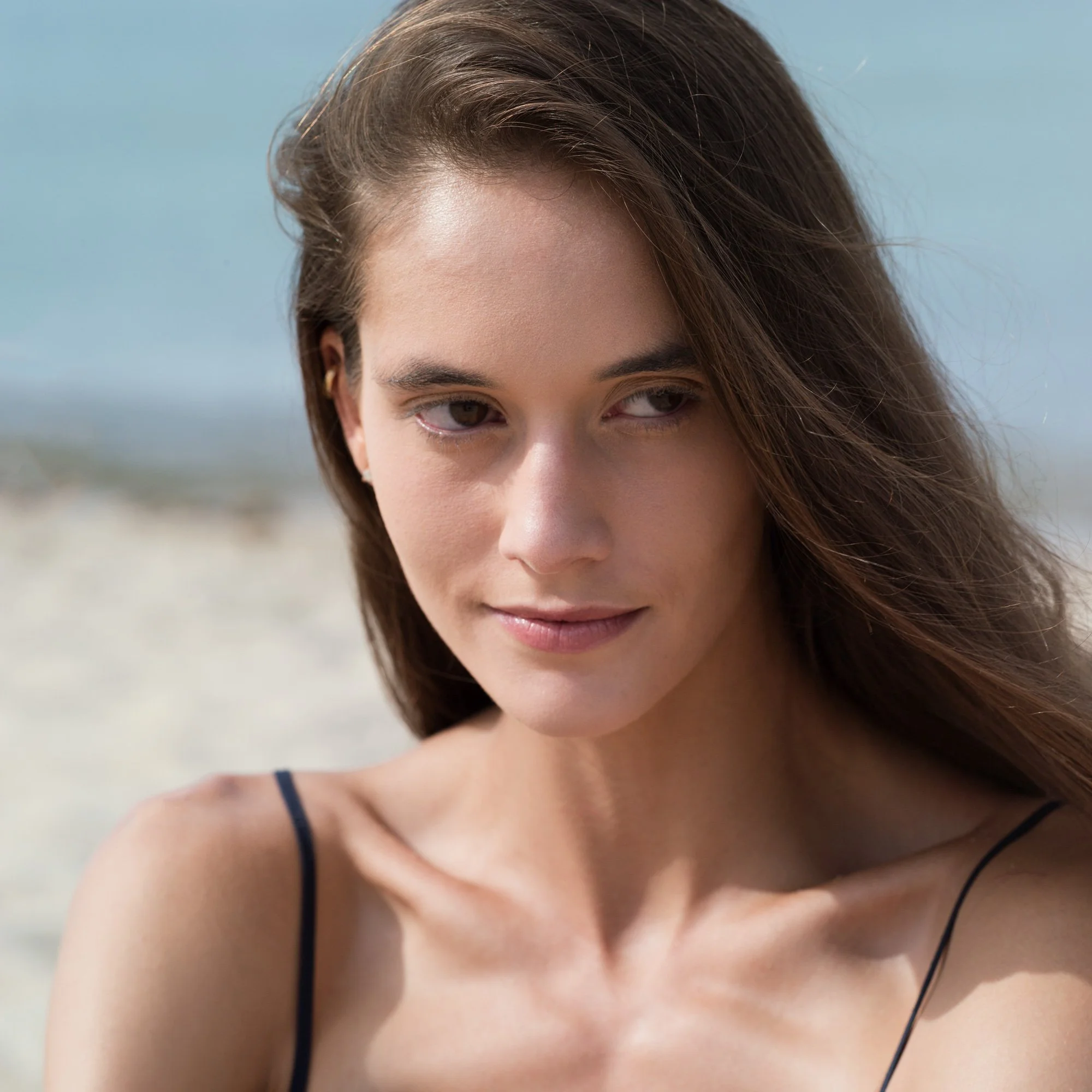 Close-up of a woman's face, showing her forehead, eyes, and part of her nose. She has brown hair, light skin, and is wearing a thin black strap on her shoulder. The background appears to be outdoors with a blurred landscape.