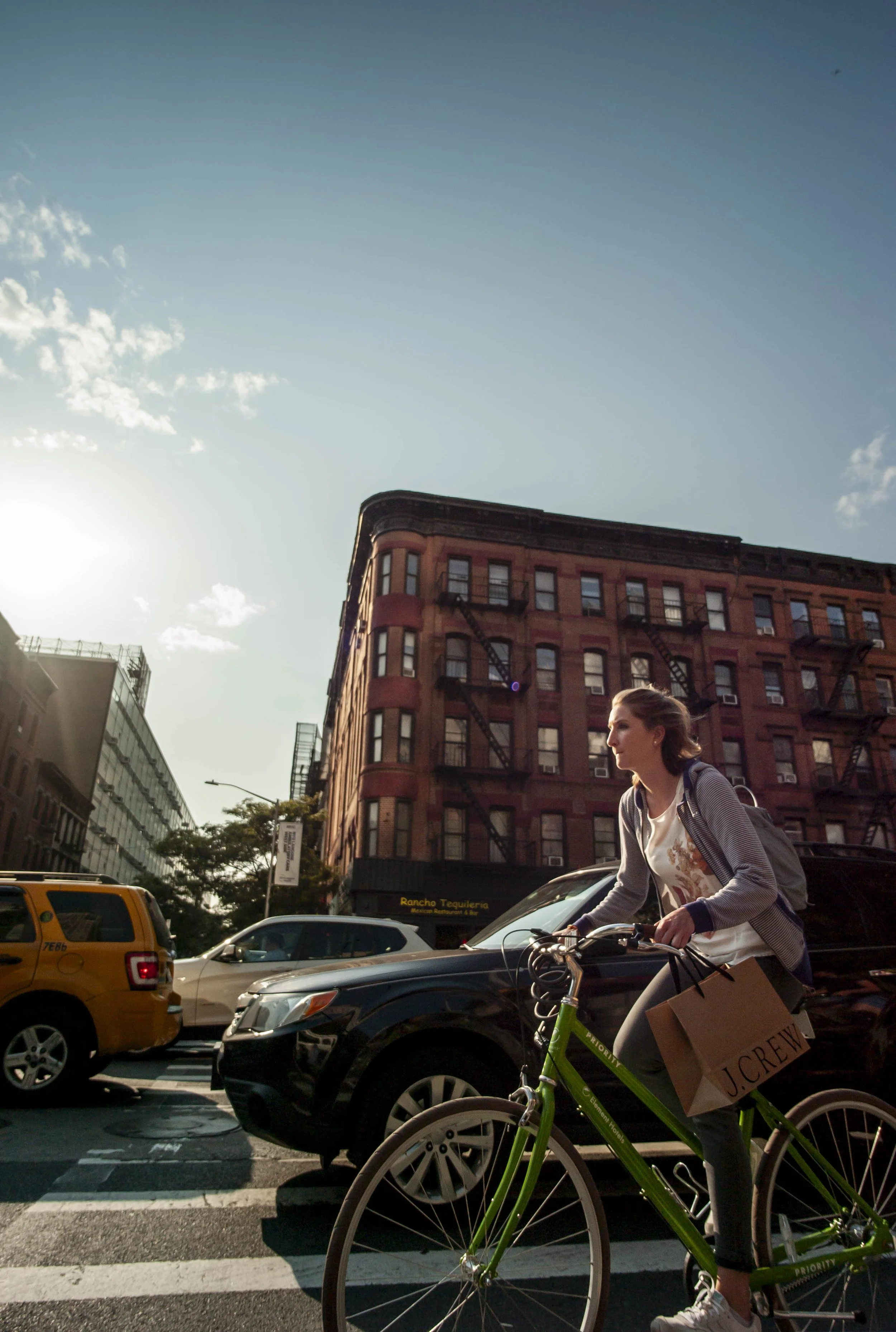 A woman riding a green bicycle across a city street with cars and tall apartment buildings under a partly cloudy sky.